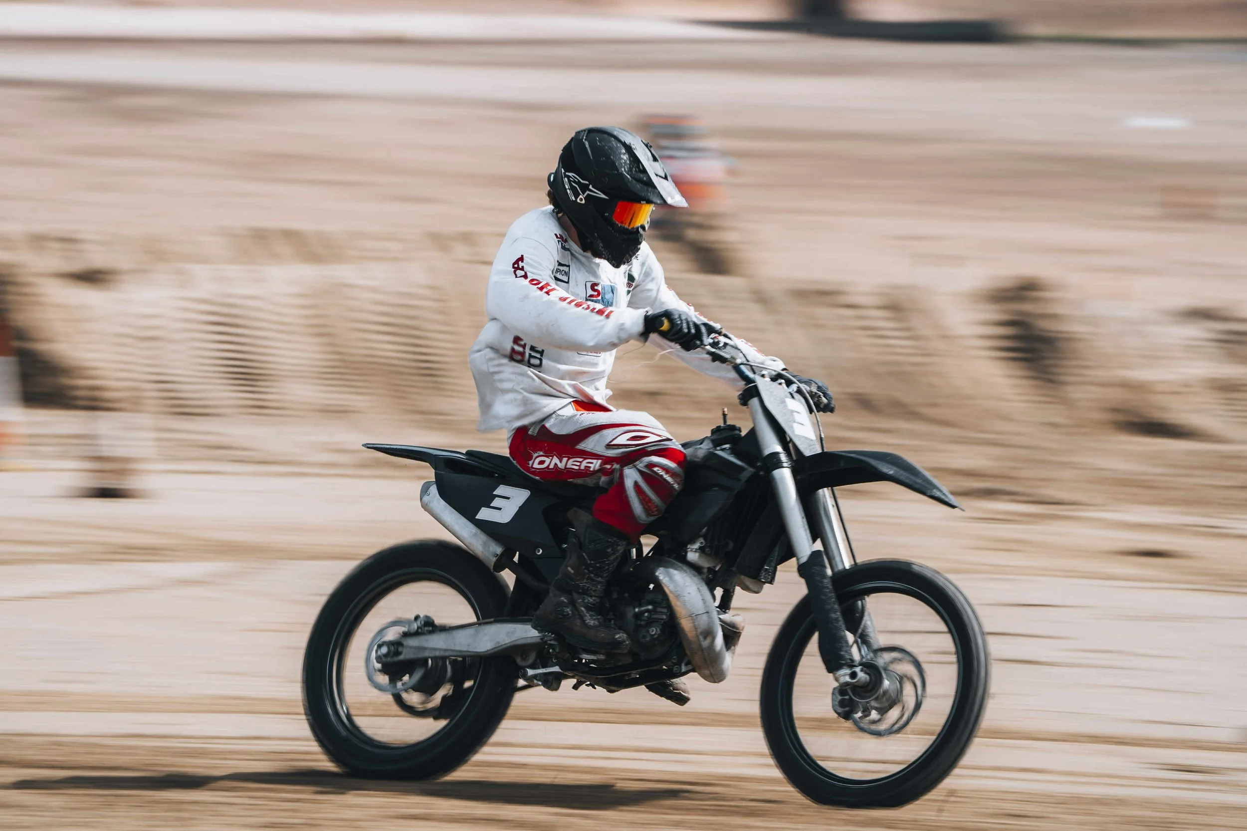 A motocross rider wearing a black helmet with a red visor, white jersey with red and black accents, and red and white pants, riding a black dirt bike with the number 3 on a sandy track.