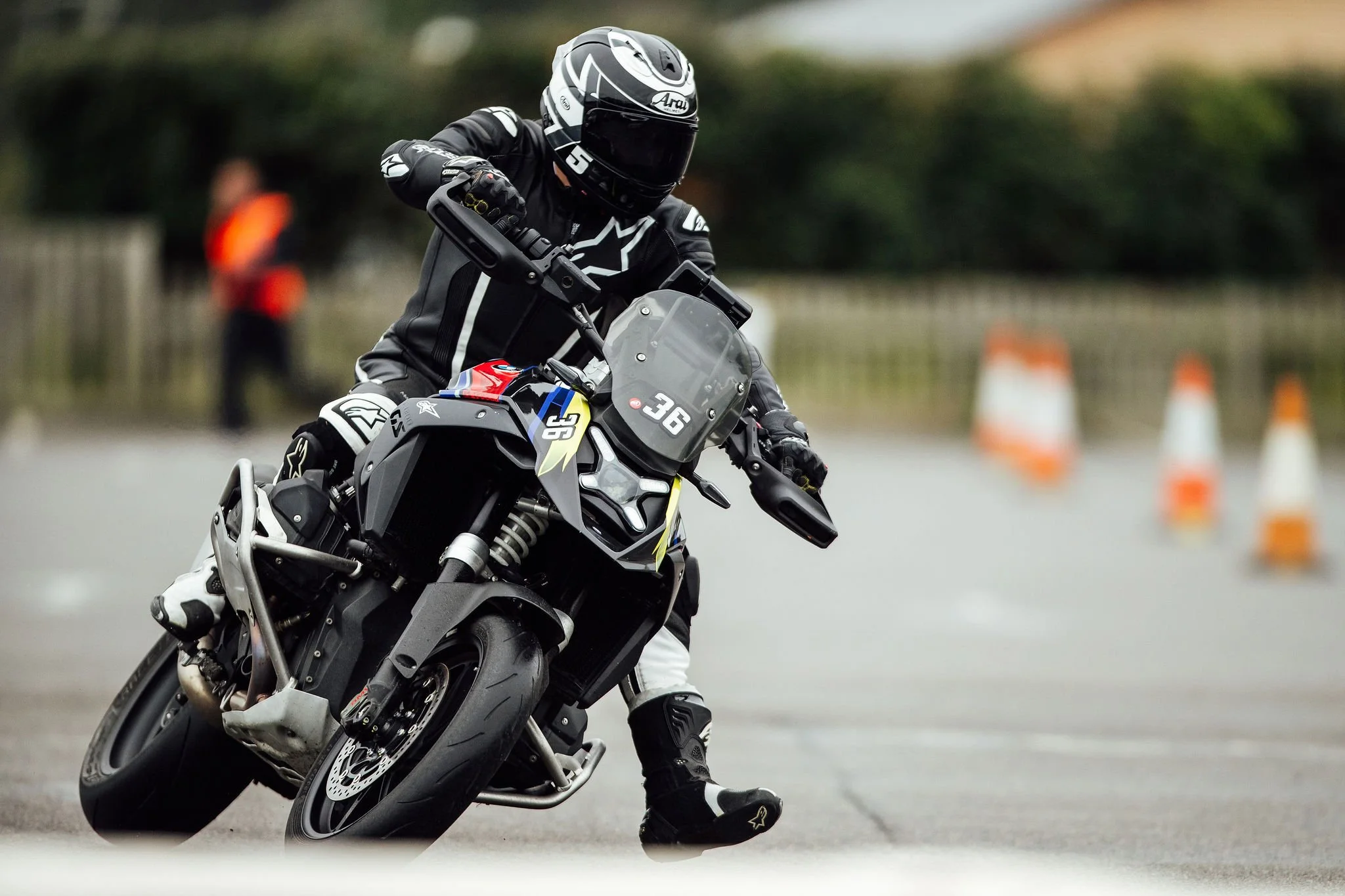 Motorcycle rider wearing black protective gear and helmet, leaning into a turn on a racing track with orange safety cones in the background.