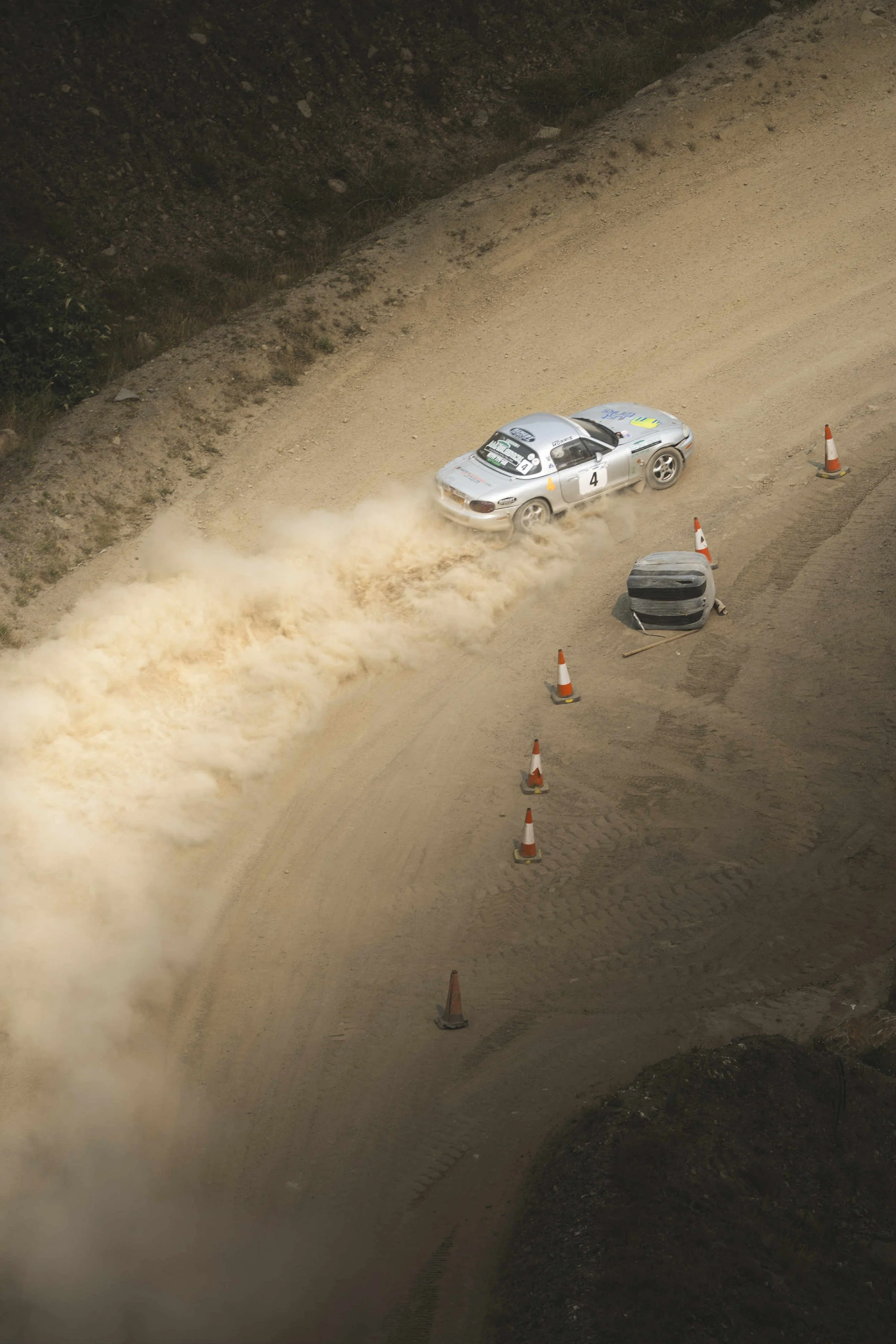 A silver race car kicking up dust on a dirt road, surrounded by orange traffic cones and barriers.