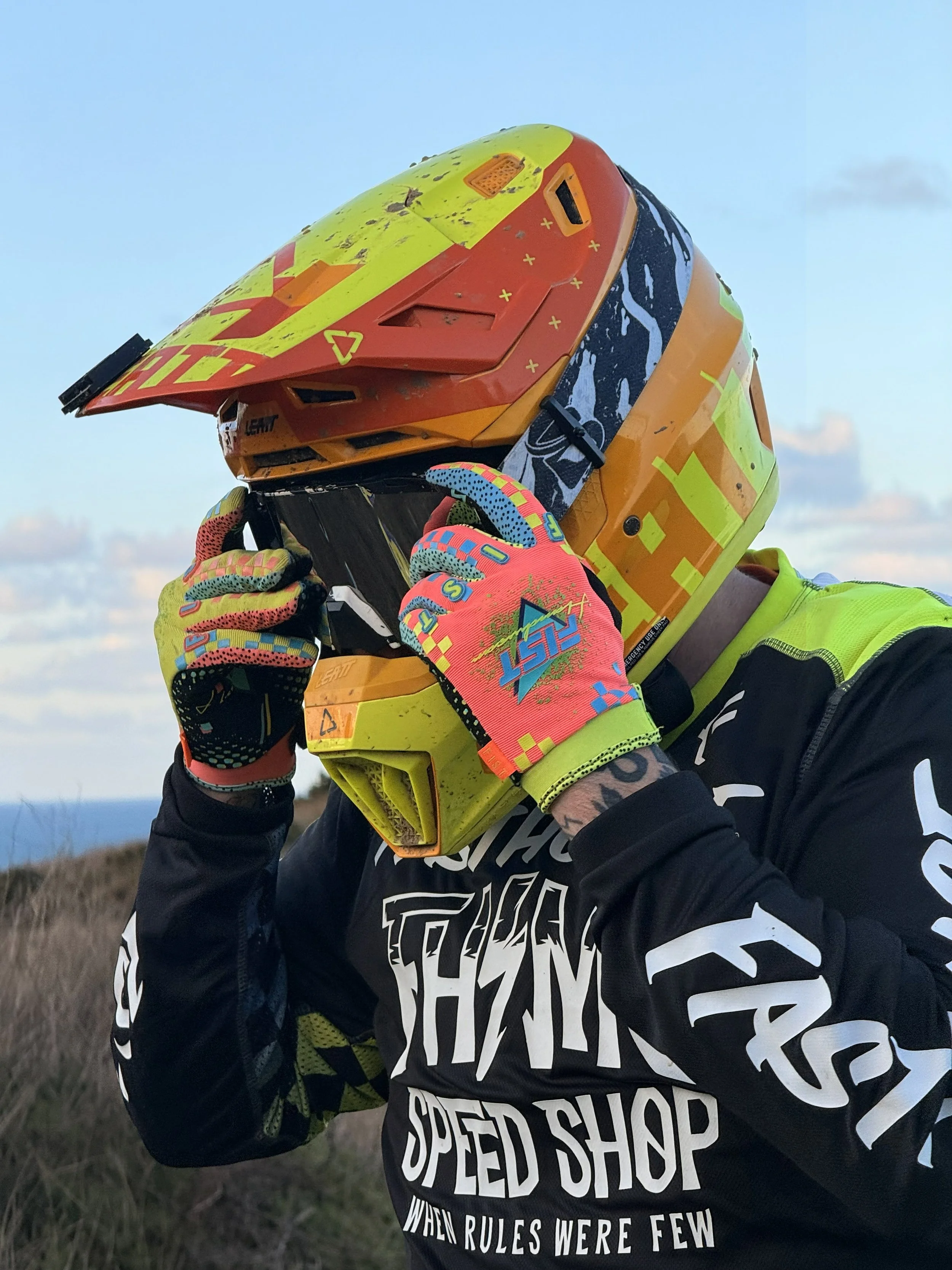 A person wearing a colorful motocross helmet, bright gloves, and a black and yellow shirt holding the helmet close to their face outdoors.