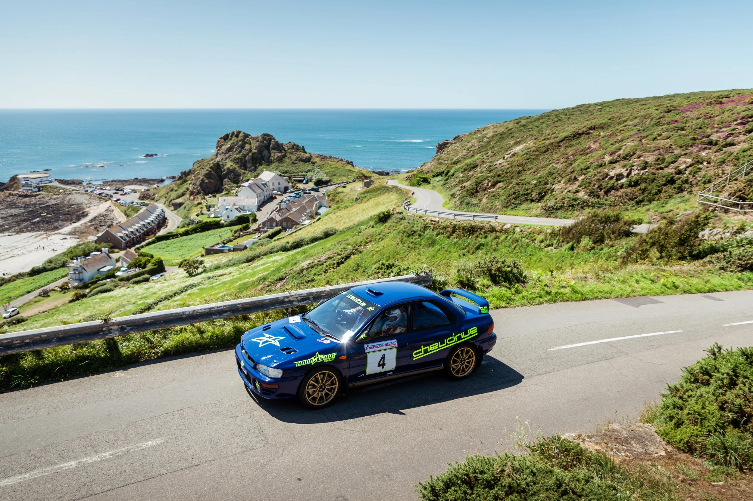 A blue rally car with the number 4 on the side driving along a coastal road with green hills, houses, and the ocean in the background.