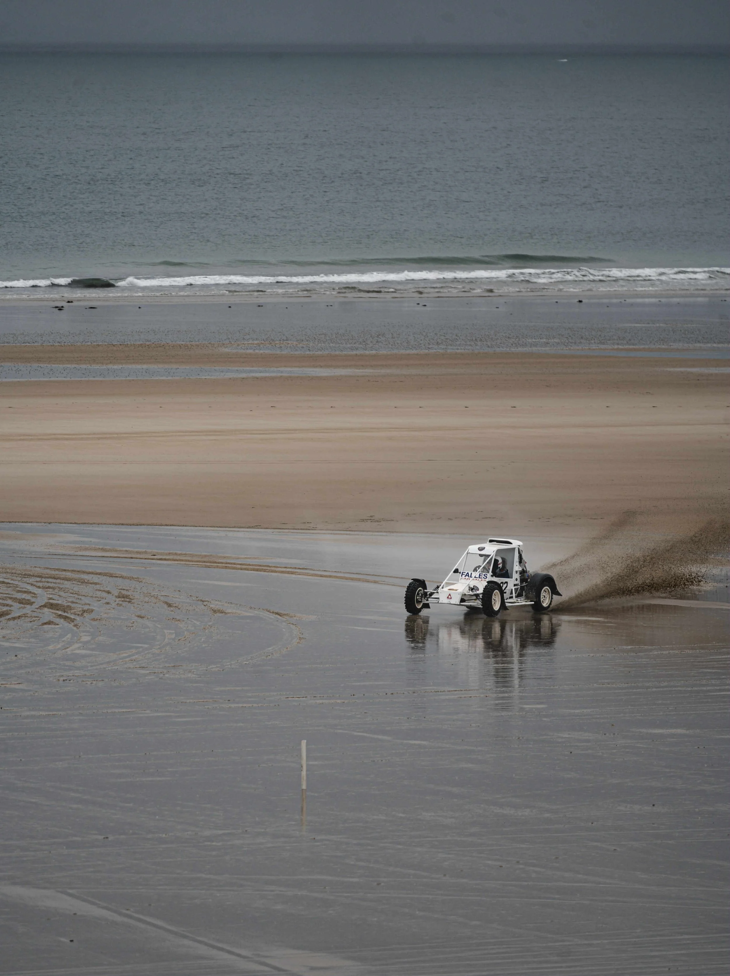 A small dune buggy driving on a sandy beach near the shoreline, with the ocean waves in the background.