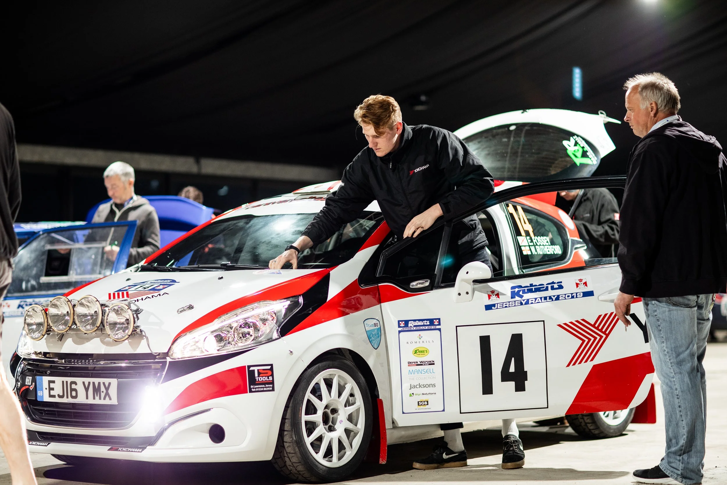Rally race car with number 14 being prepared in a garage, with team members working on the car.