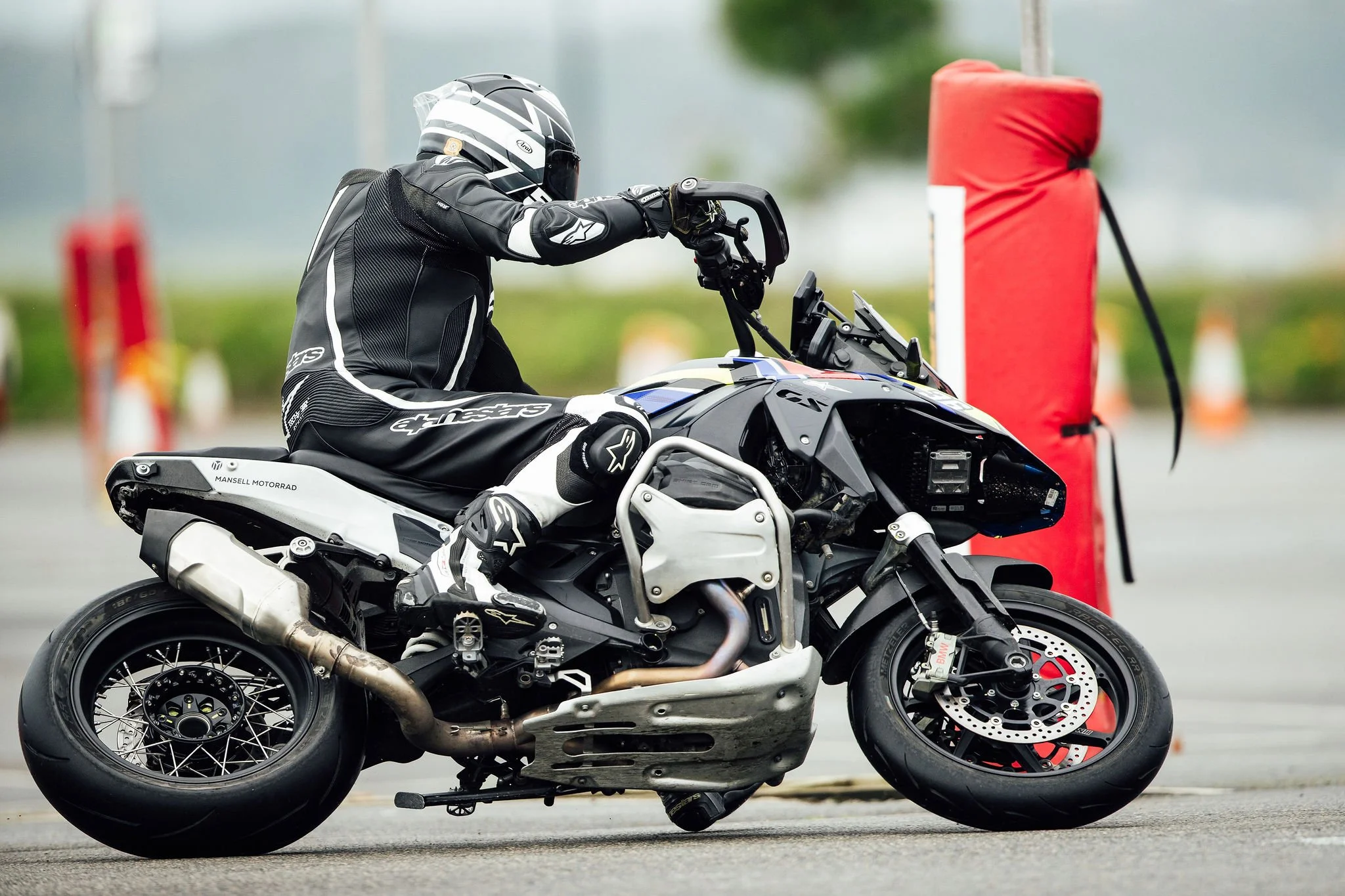A person in motorcycle gear riding a black and white motorcycle on a paved surface, with an orange safety cone and a red padded obstacle in the background.