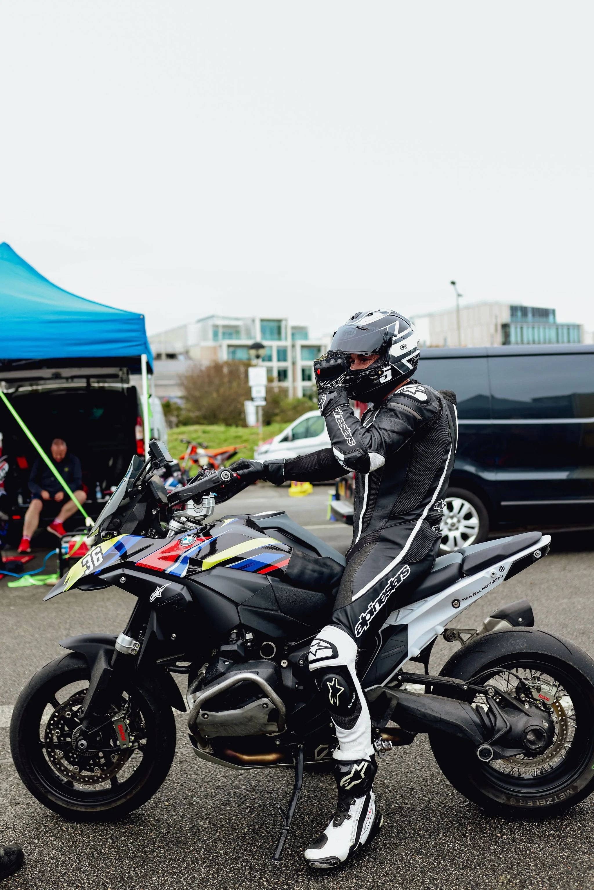 A motorcyclist dressed in racing gear with a helmet and suit, sitting on a black motorcycle with a colorful BMW design, in a paddock area with tents and vehicles.