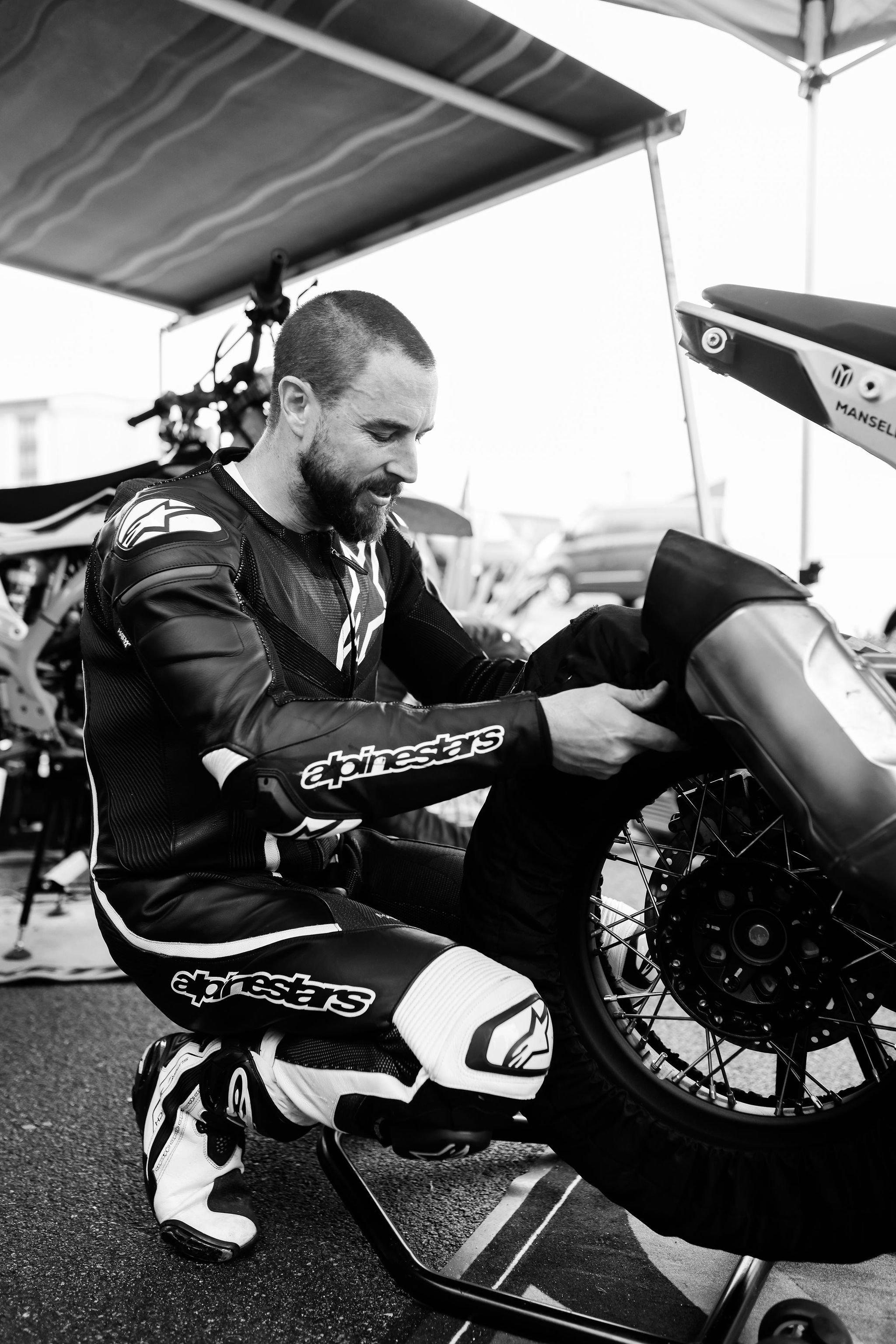 A man in motorcycle racing gear working on a dirt bike under a tent