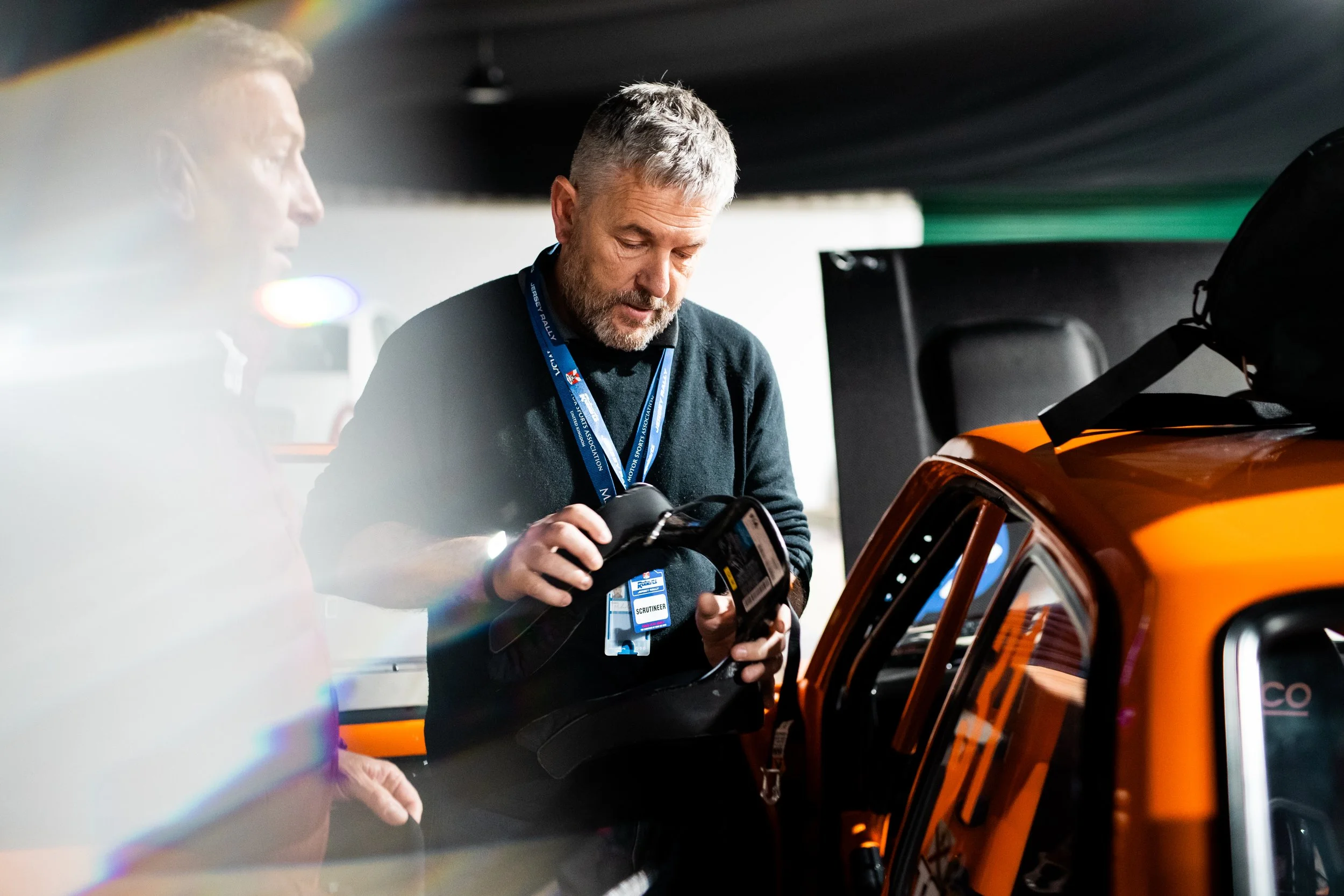 A man with gray hair and beard, wearing a black sweater and a lanyard, is examining a device while standing next to an orange race car inside a garage.