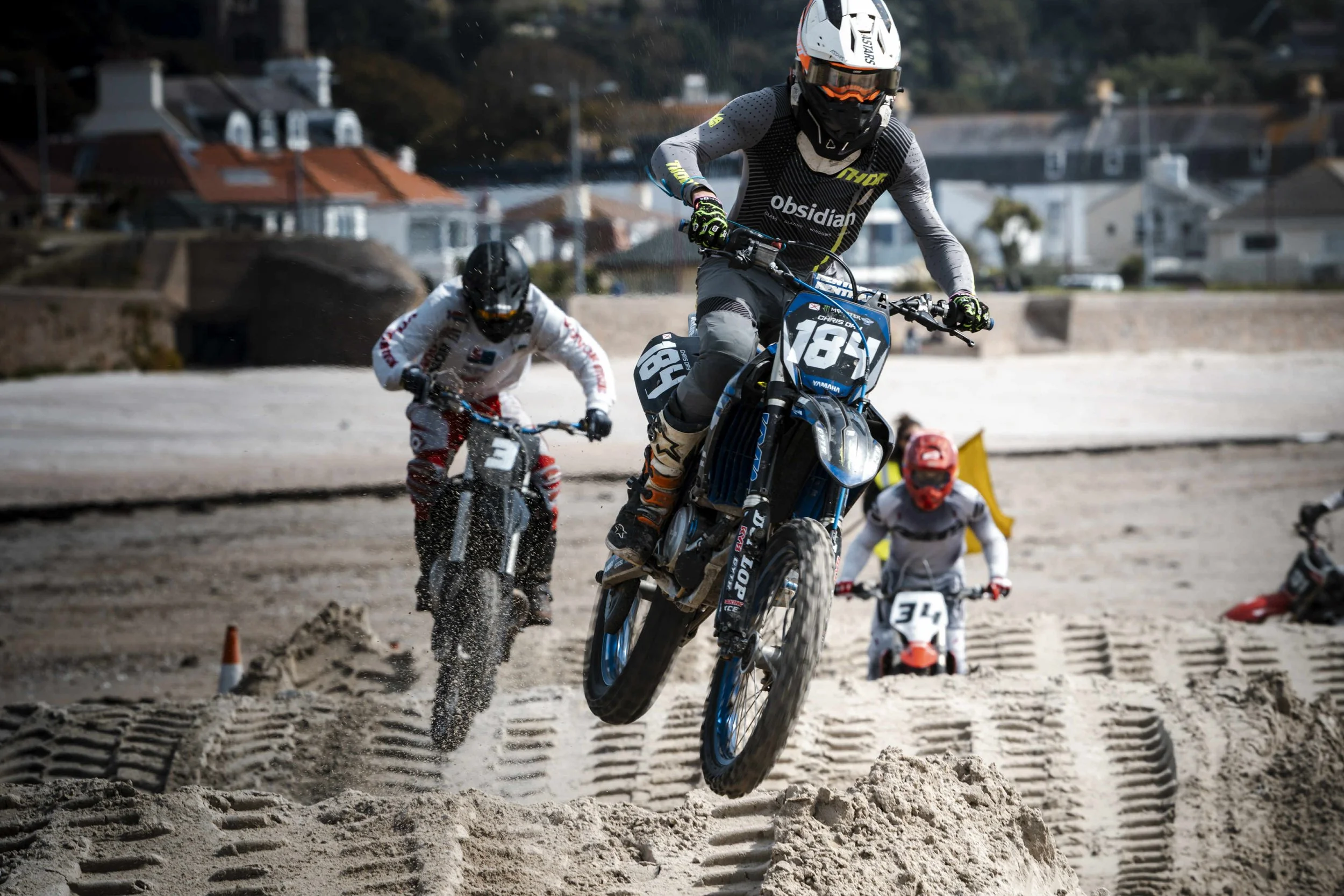 Motocross racers riding on sandy terrain with three riders jumping over a sand mound, houses in the background.