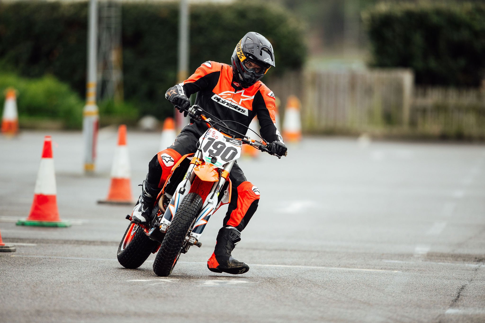 Motorcycle rider navigating a course marked with orange cones, wearing a black and orange racing suit and helmet, on an asphalt surface.