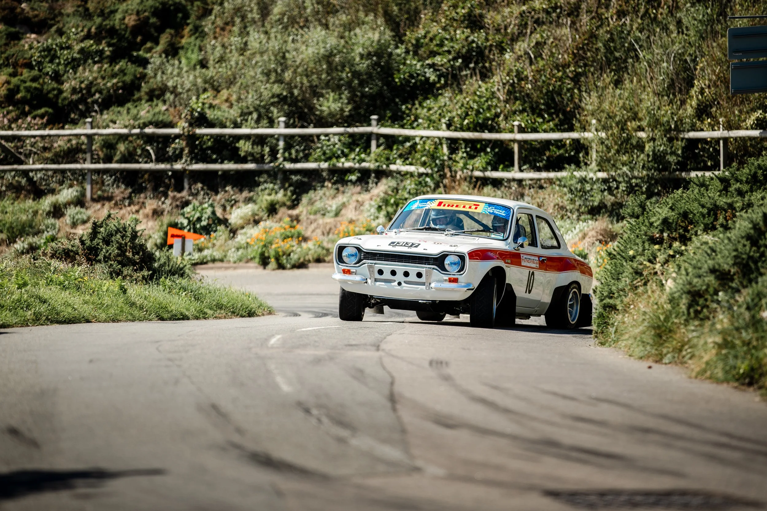 A vintage rally car with a white body and red stripe, number 10, navigating a winding mountain road surrounded by lush green bushes and trees.