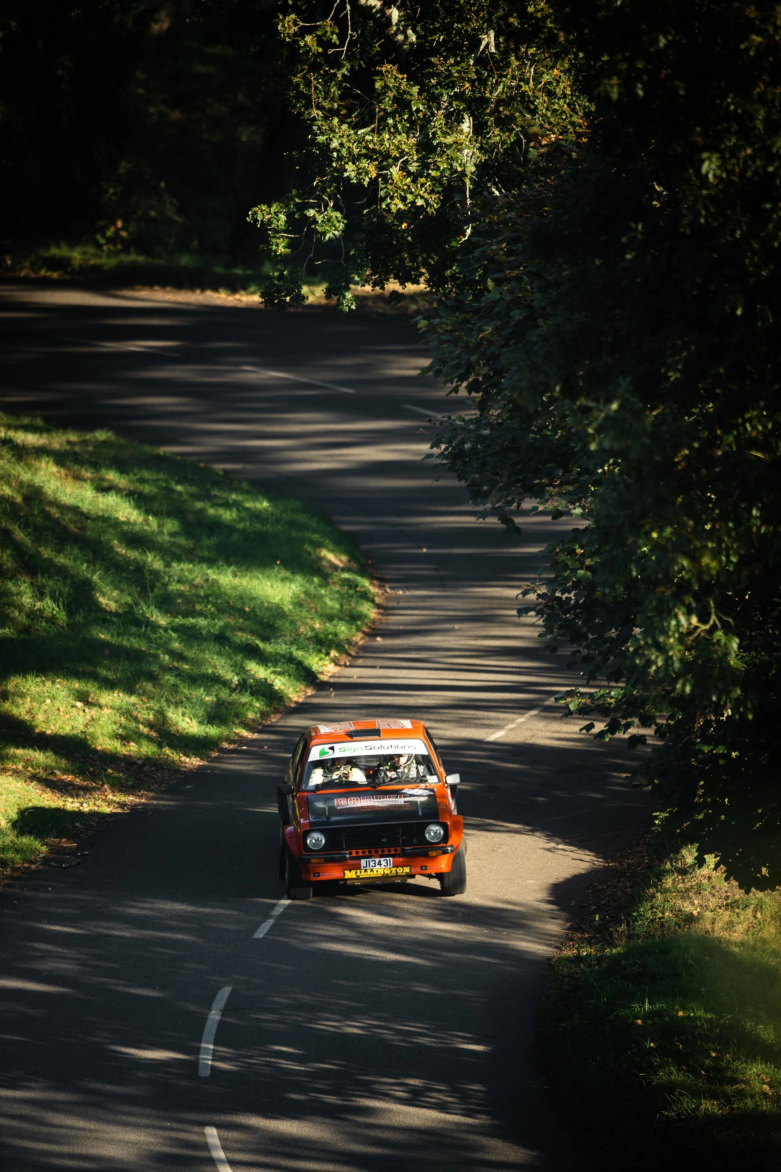 A vintage rally car driving on a tree-lined rural road during the daytime.