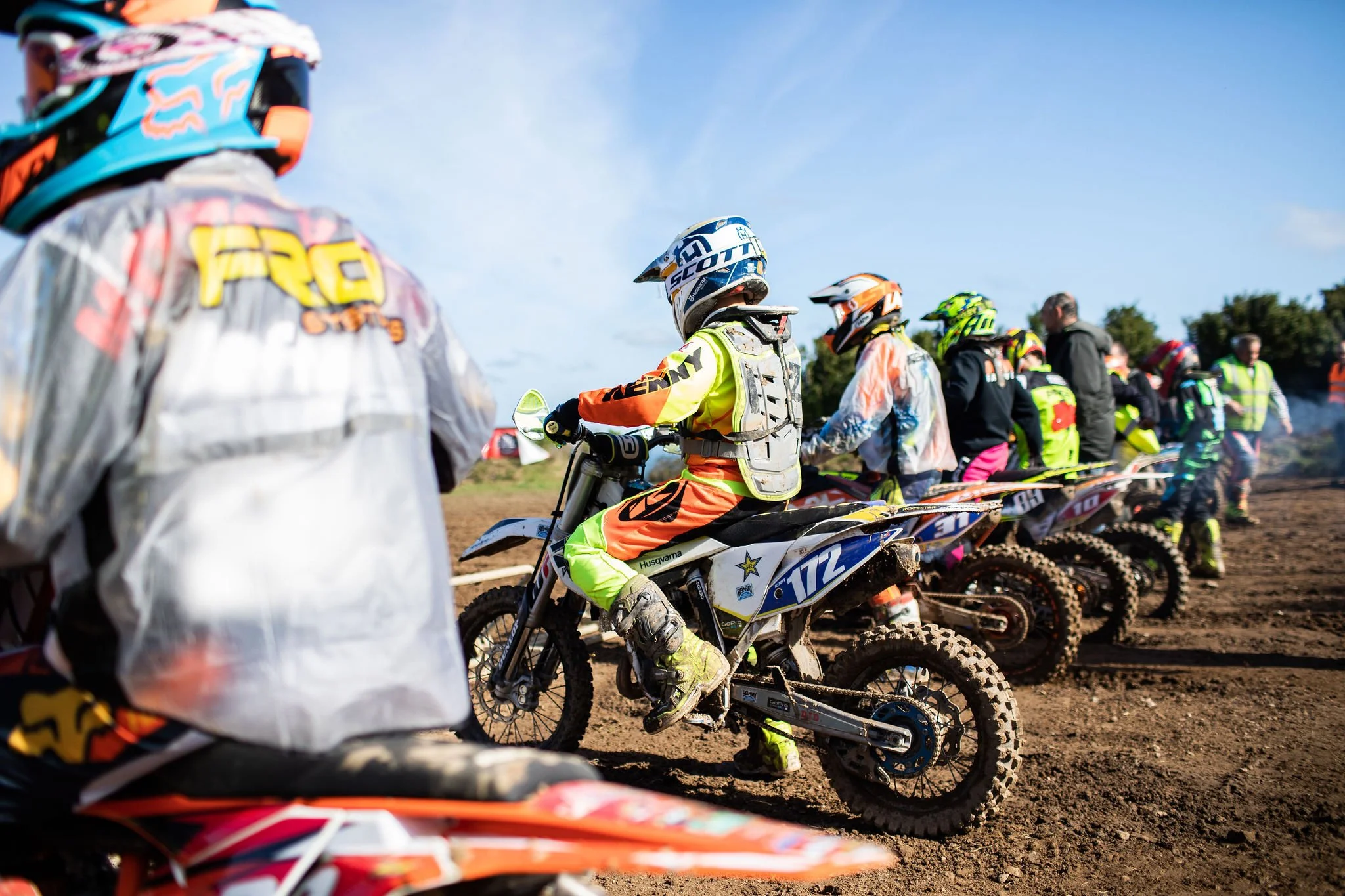 Motocross riders lined up on dirt track with helmets and gear, preparing for race, with a clear blue sky and trees in the background.