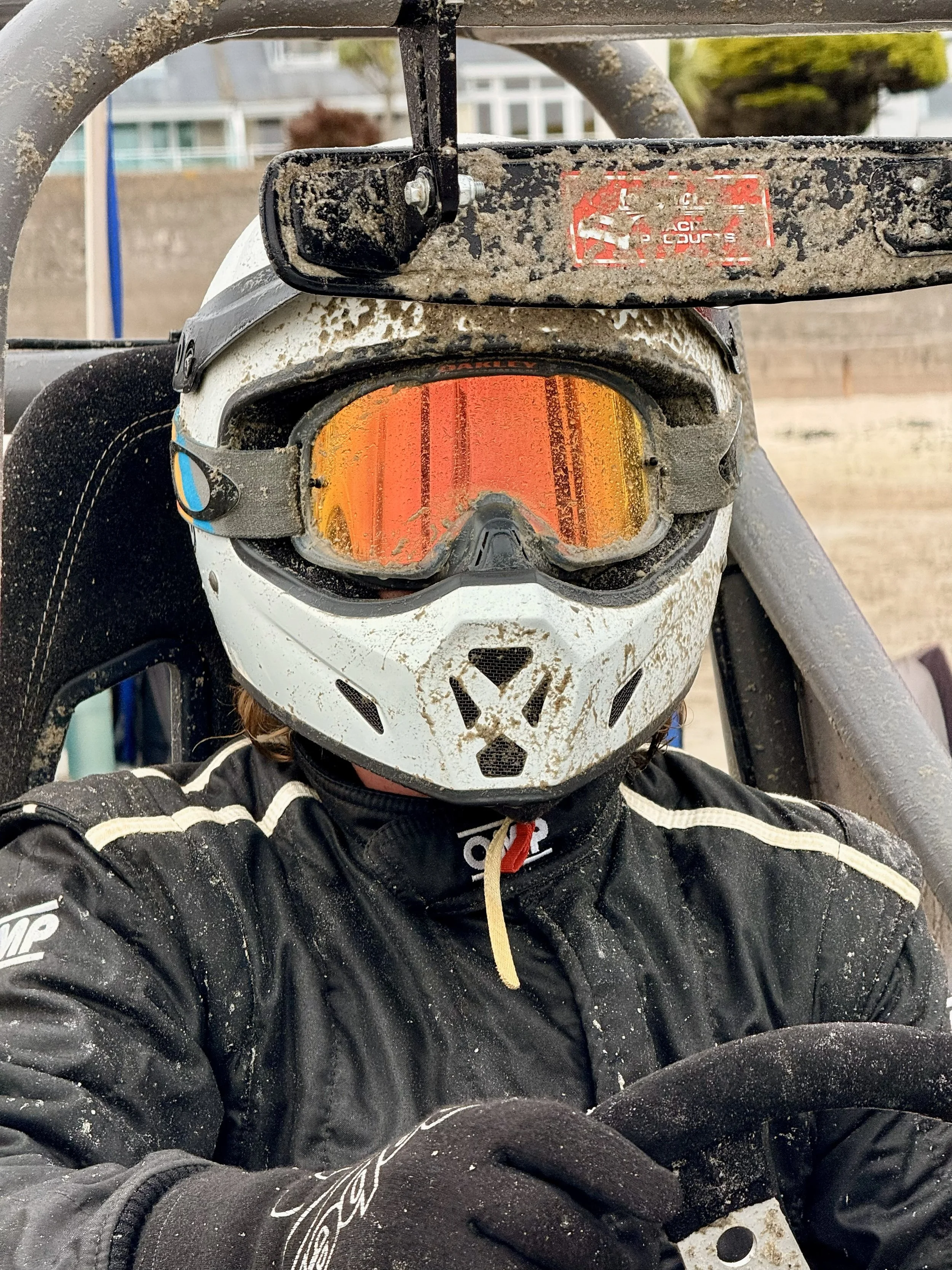 A person wearing a white motocross helmet with a black visor, orange tinted goggles, black gloves, and a black racing suit sitting in a dirt-service vehicle, with muddy gear and a blurred outdoor background.