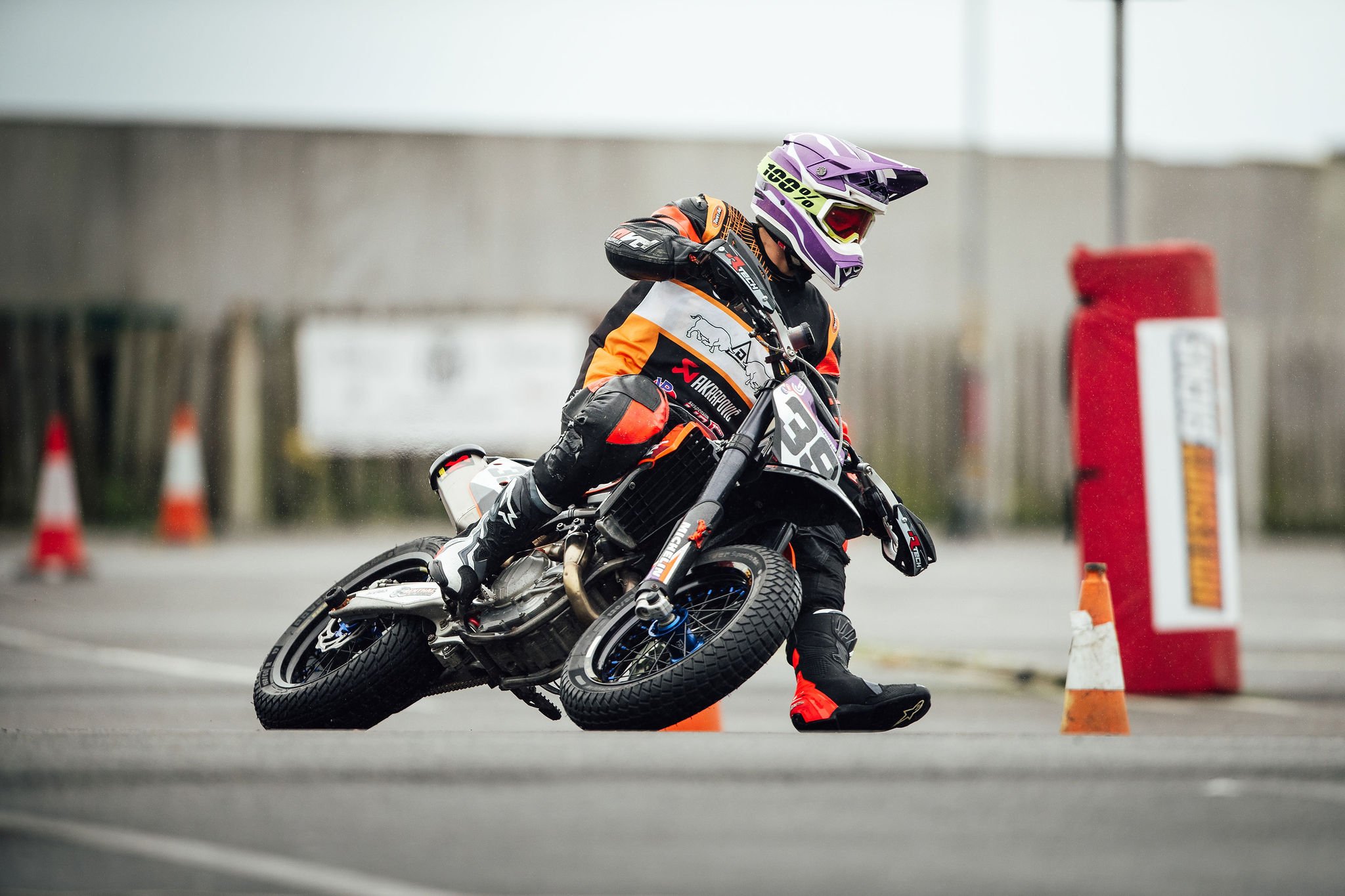 Motorcyclist in racing gear leaning into a turn during a ride on a paved surface with orange traffic cones and a red barrier in the background.
