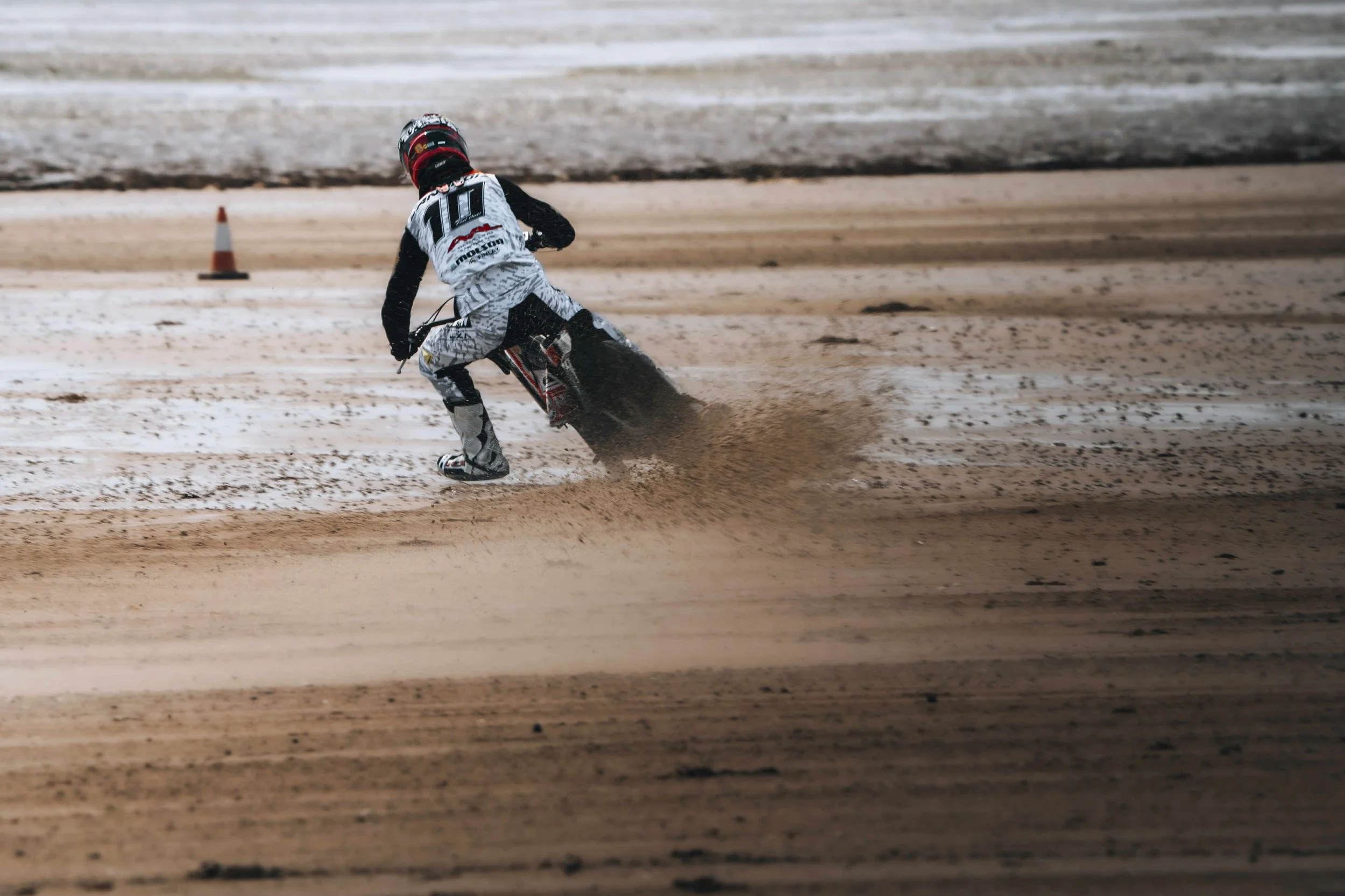 Speedway rider in racing gear on dirt track, leaning into turn, kicking up dust.