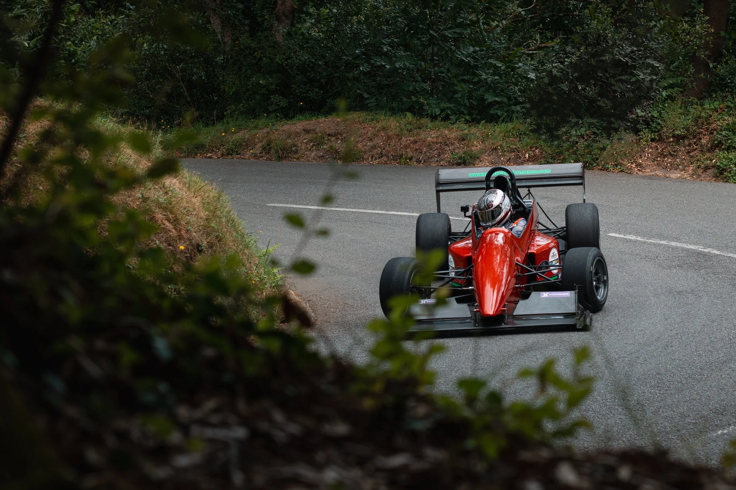 A red open-wheel race car speeds around a curve on a winding mountain road, with lush green foliage in the background.