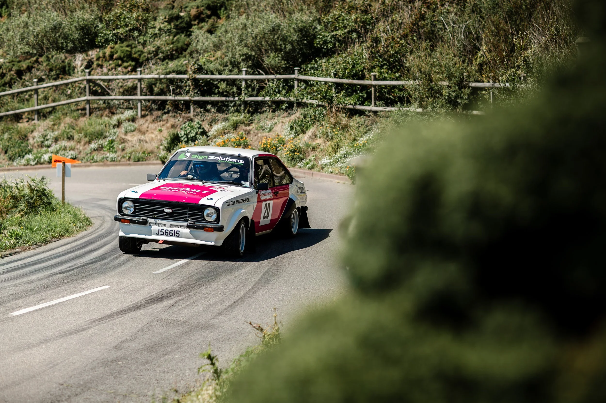 A vintage race car with the number 20 on its side and a license plate J66I5 navigating a sharp turn on a mountain road, partially obscured by bushes in a scenic countryside setting.