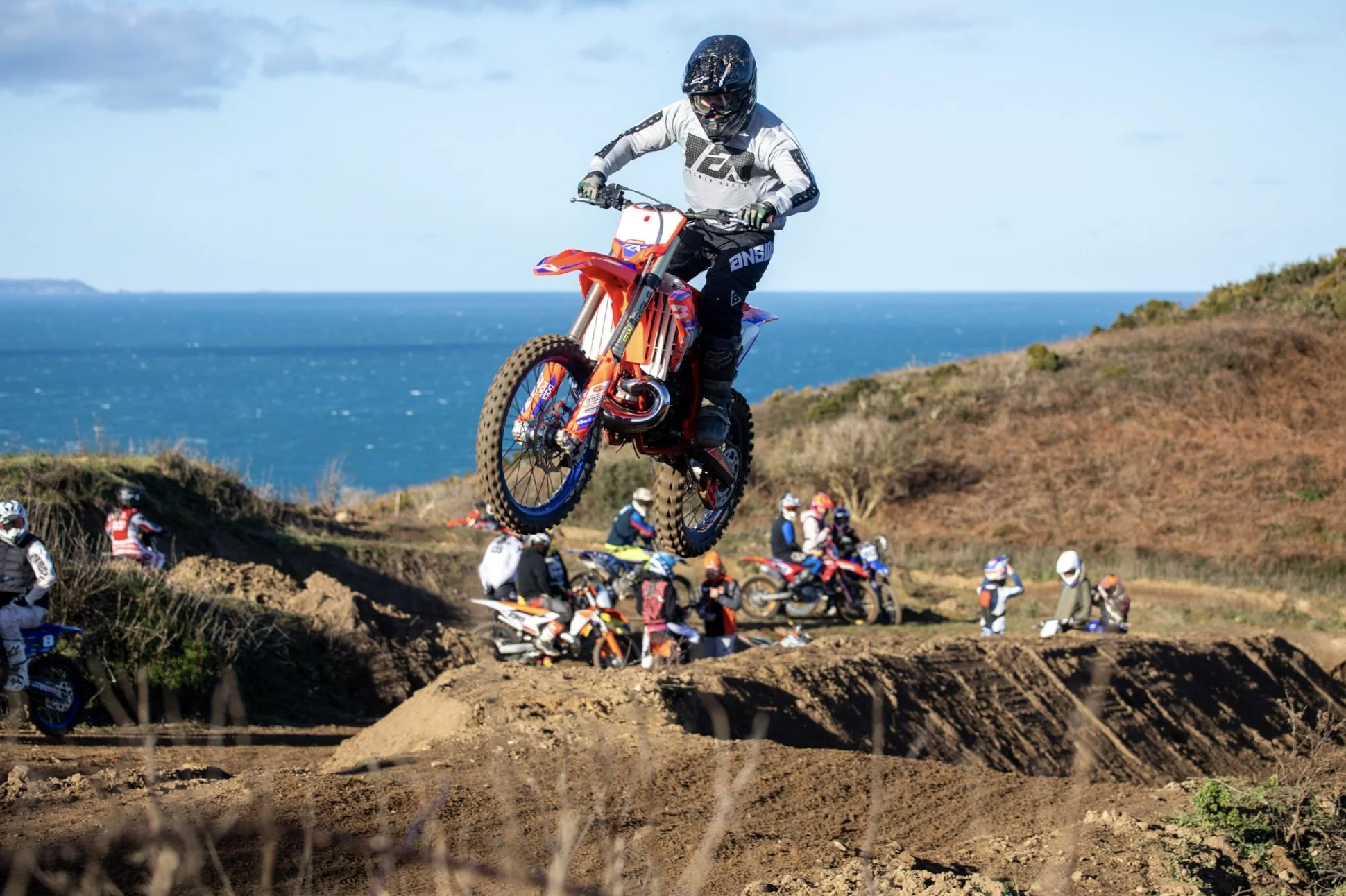 A dirt bike rider in mid-air jump, overlooking a group of other riders and spectators on a dirt track near the ocean.