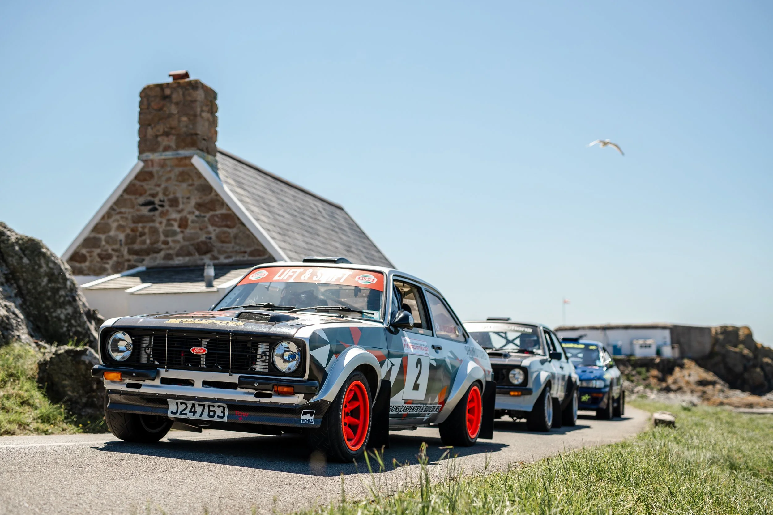 A line of rally cars, including a black and gray car with red wheels and racing decals, driving on a narrow road near a stone cottage with a chimney, under a clear blue sky with a seagull flying overhead.