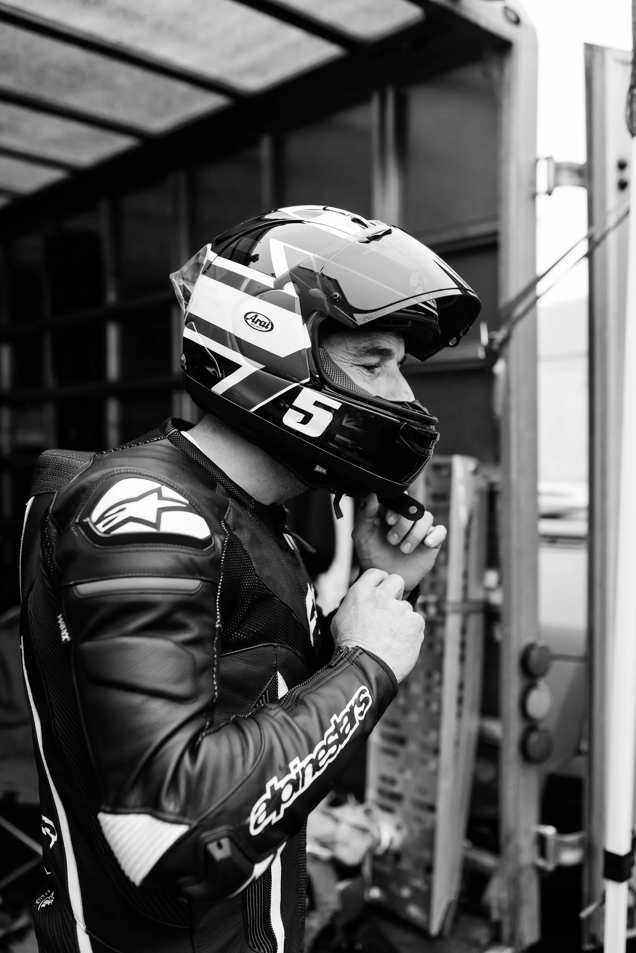 A race car driver wearing a racing suit with Alpinestars branding and an Arai helmet, preparing for a race in the garage.