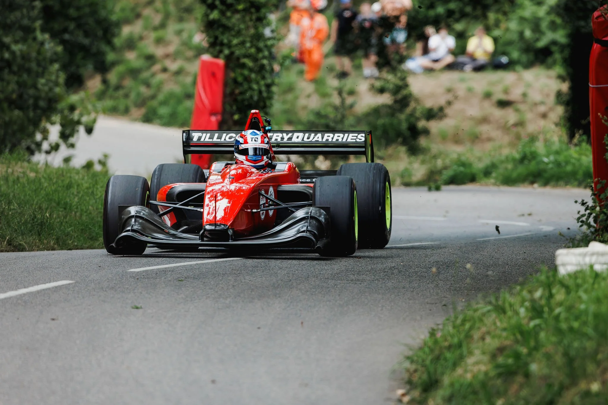 A red Formula racing car with the driver wearing a blue, red, and white helmet, moving on a narrow road with trees and bushes on the sides. People are sitting on the hill in the background, watching the race.