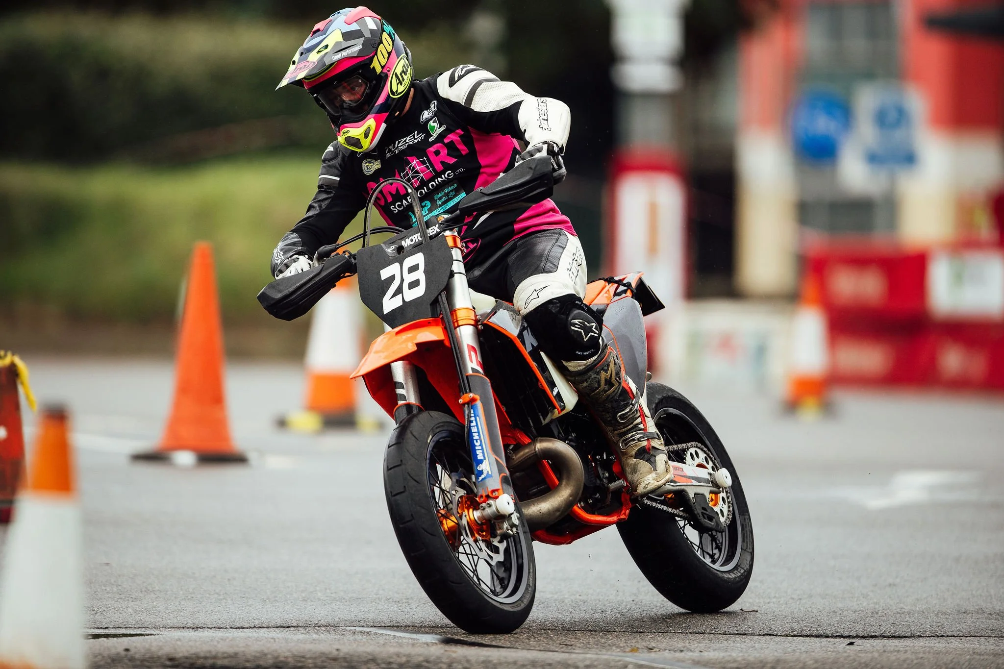 Motorcycle racer wearing a helmet and protective gear, riding an orange and black motorcycle through a slalom course with orange traffic cones.