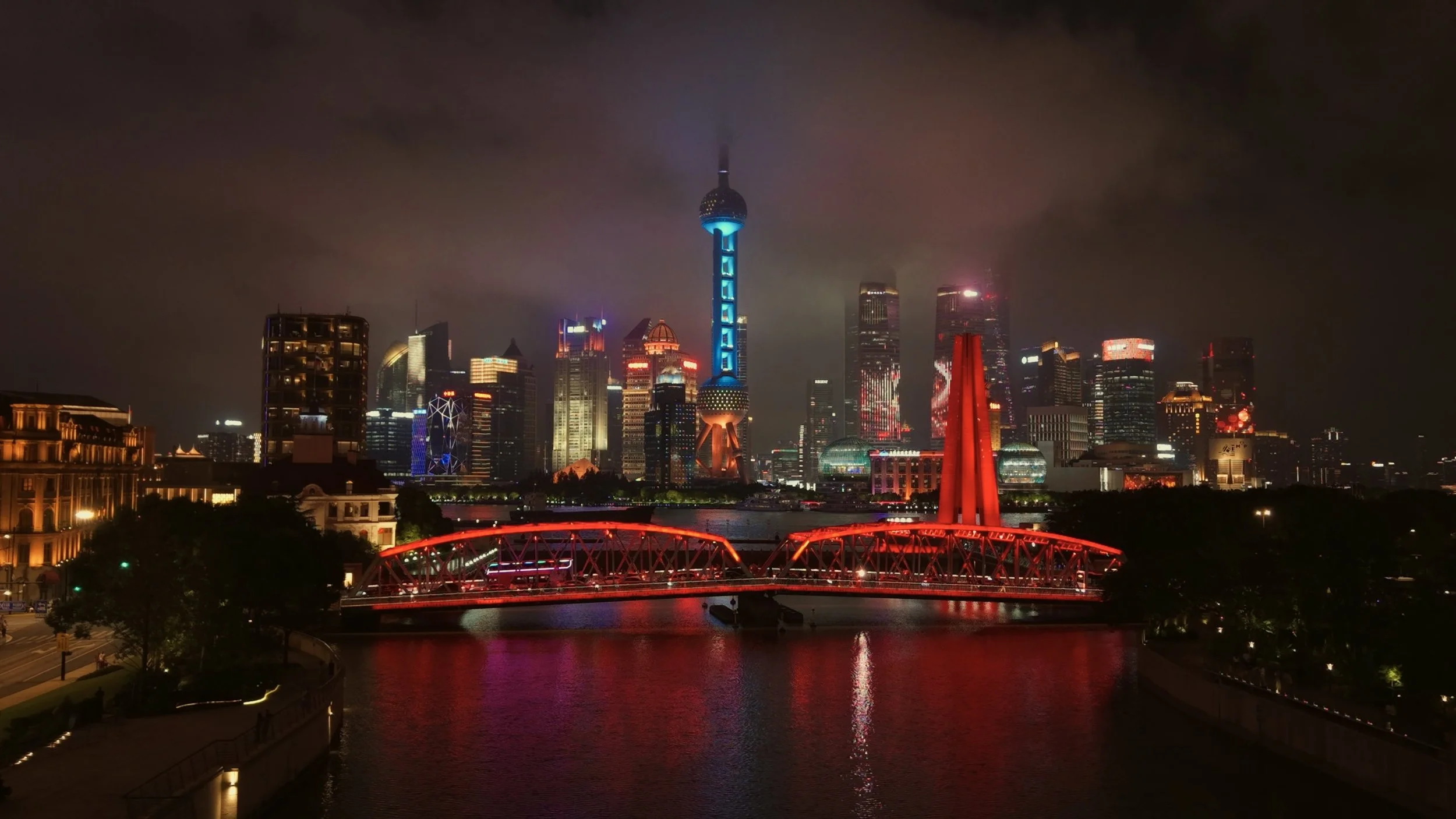 Night view of Shanghai skyline with illuminated skyscrapers, the Oriental Pearl Tower, and a red bridge over the river.