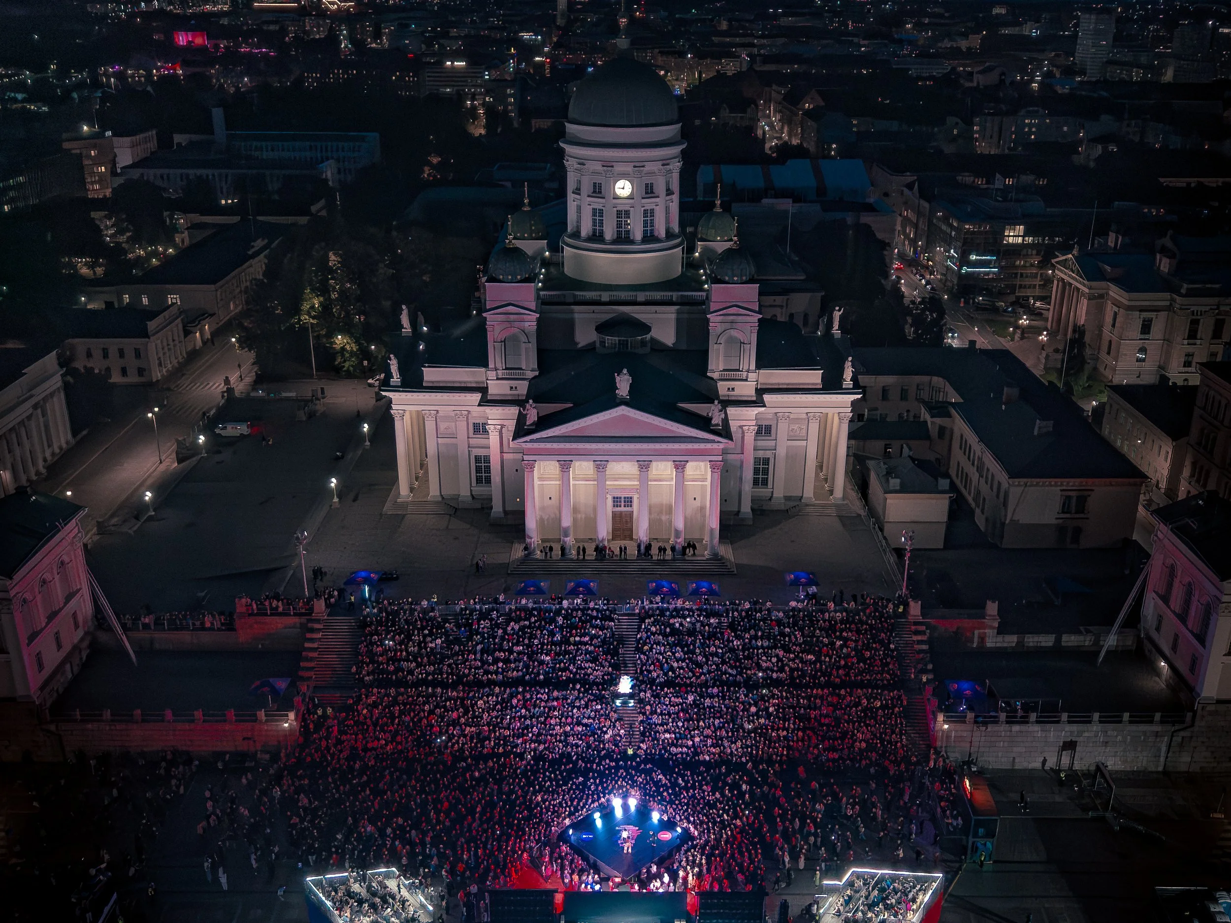 Nighttime aerial view of a large assembly of people gathered in front of a historic building with a domed clock tower, including pink lighting, across a cityscape with illuminated streets and buildings in the background.
