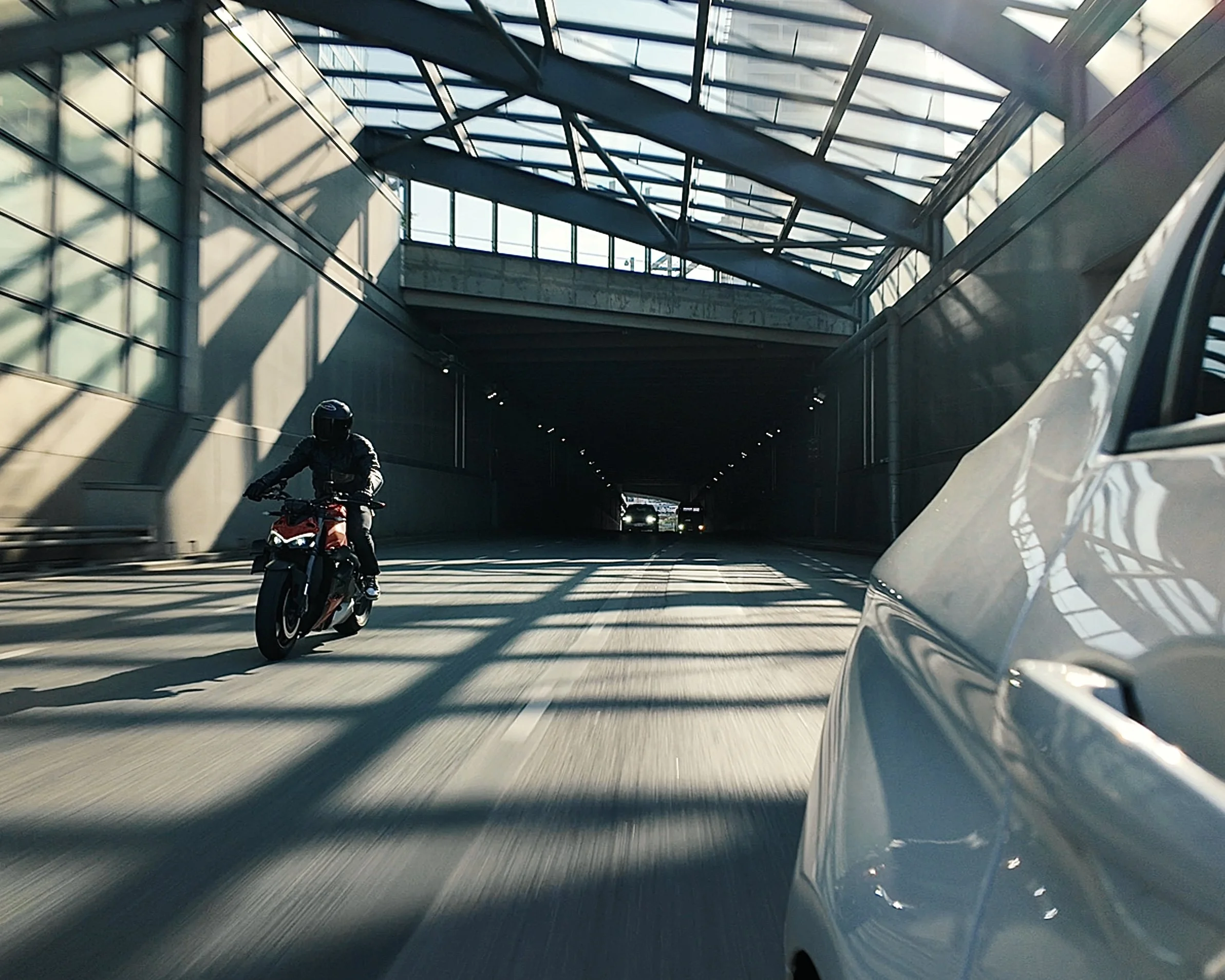 A motorcycle rider and a car are traveling through a tunnel with glass walls and ceiling, with sunlight casting shadows inside.