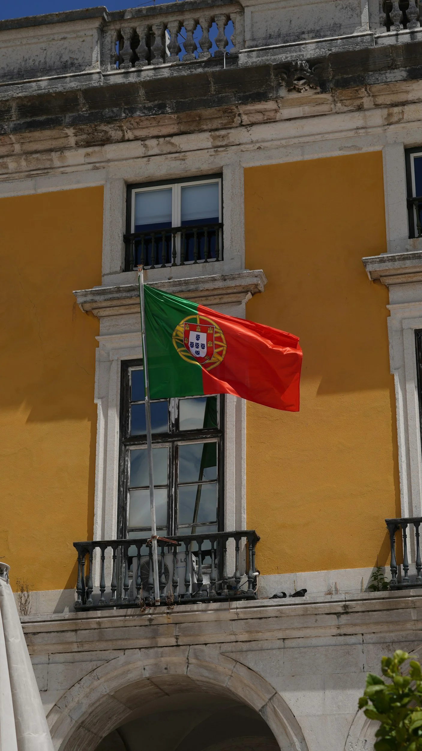 A Portuguese flag hanging on a balcony of a yellow building with white stone details and an archway.