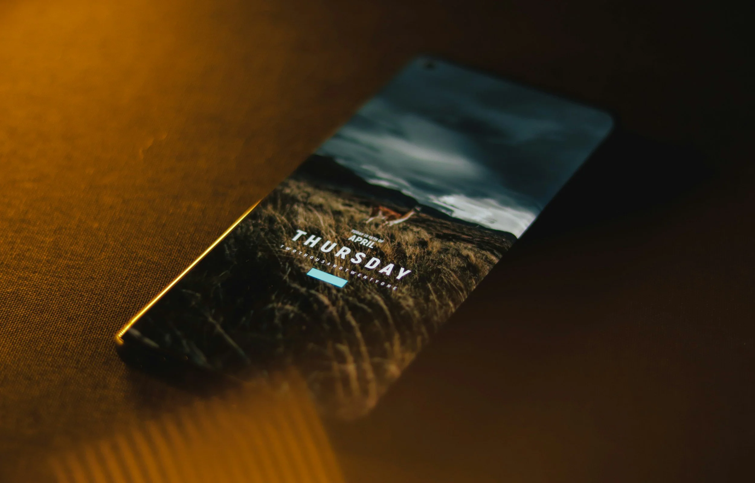 Smartphone lying on a dark surface displaying a weather forecast for Thursday, with a landscape background and cloudy sky.