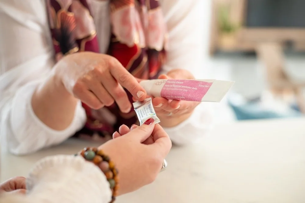 Patient holding a small white packet, while Dr Lucy Mather opens a pink and white tube of cream or lotion in a medical clinic.