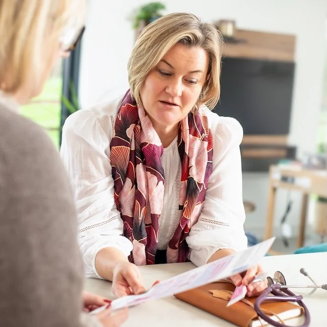 Dr Lucy Mather wearing a colourful scarf sitting at a desk, showing a document handed to a patient in a medical clinic.
