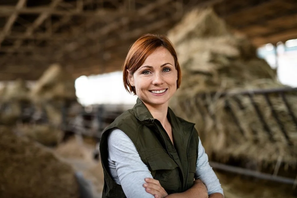A woman with short red hair smiling, standing in a barn with a cow in the background.