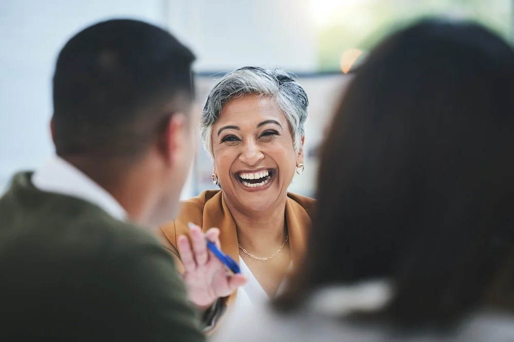 A woman with short gray hair laughing and engaging with two other people in a social setting.