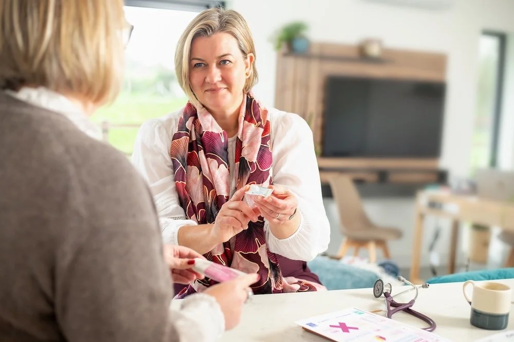 Dr Lucy Mather wearing a patterned scarf discussing medication with a patient in a medical clinic.