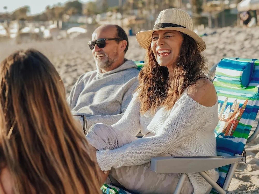 People sitting on a beach, smiling and talking. One woman wearing a straw hat and white sweater, two men with sunglasses, relaxed by the shoreline.