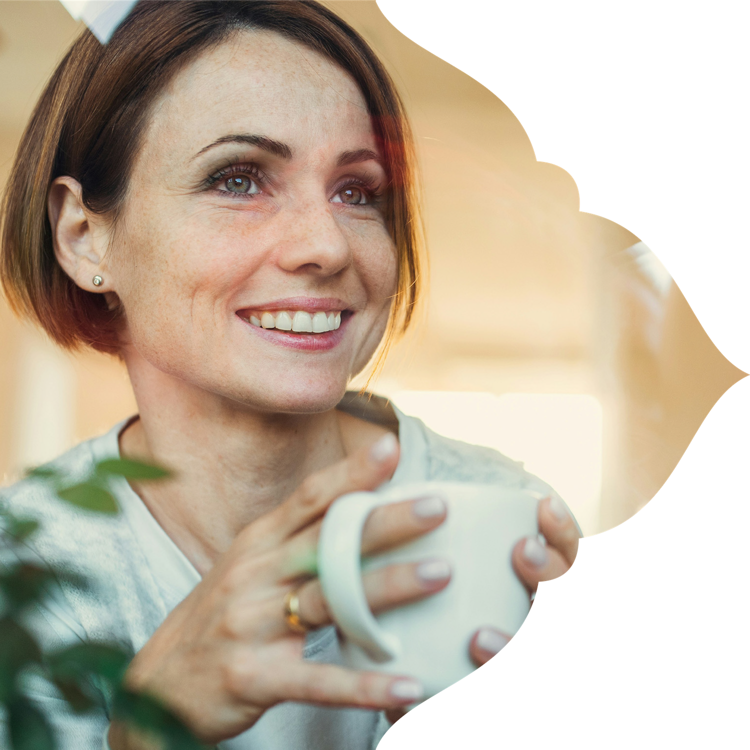 A woman with short red hair smiling while holding a white mug, sitting near a window with warm lighting.