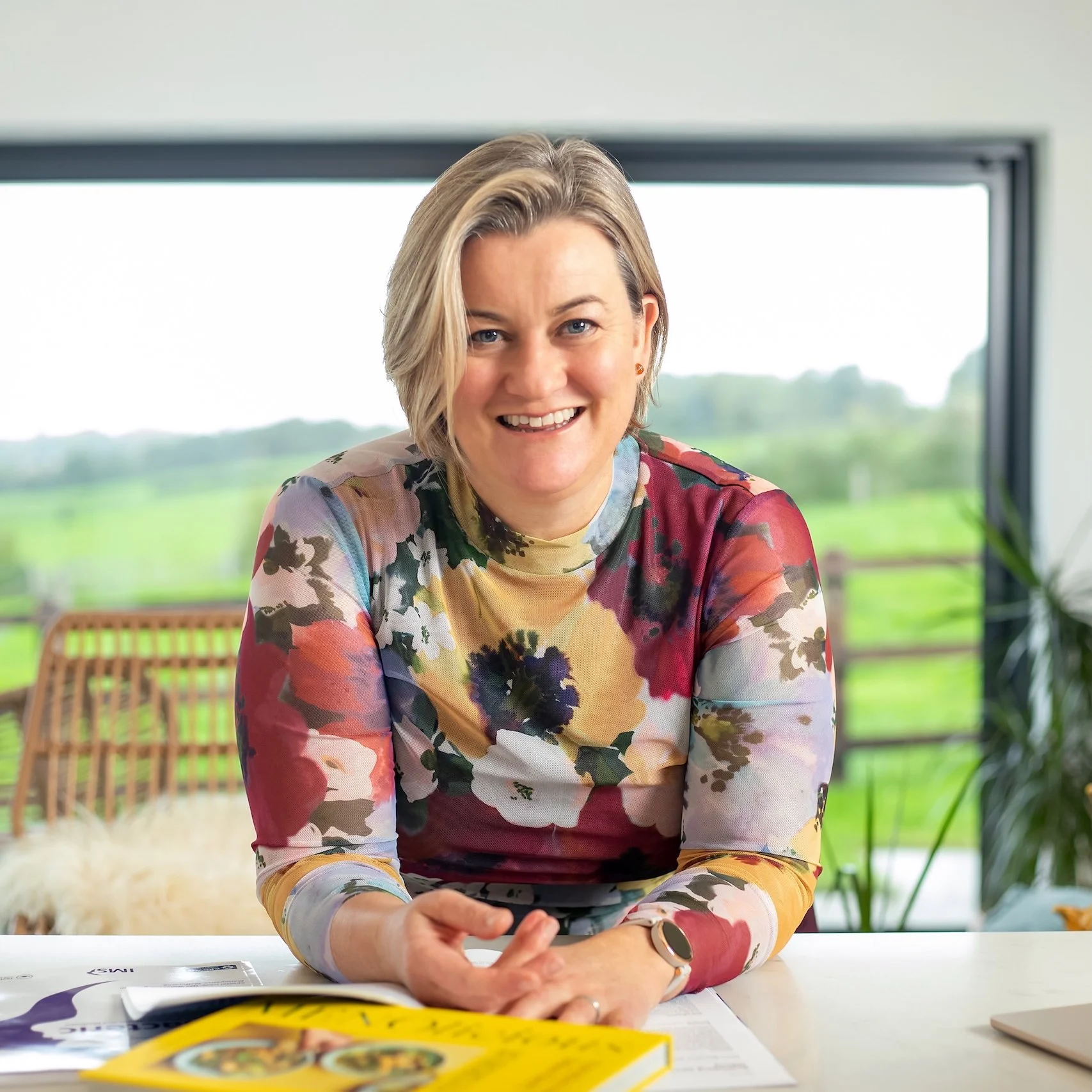 Dr Lucy Mather wearing a colourful floral blouse, sitting at a table with papers and a yellow book, smiling at the camera with a green outdoor landscape in the background.