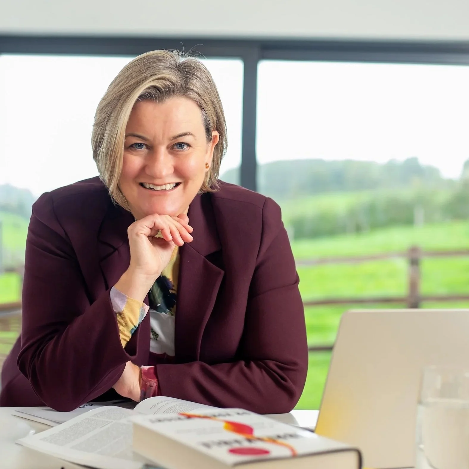 Dr Lucy Mather wearing a burgundy blazer, is smiling at the camera while sitting at a table in a bright room with large windows overlooking a green outdoor landscape, with a book and a laptop in front of her.