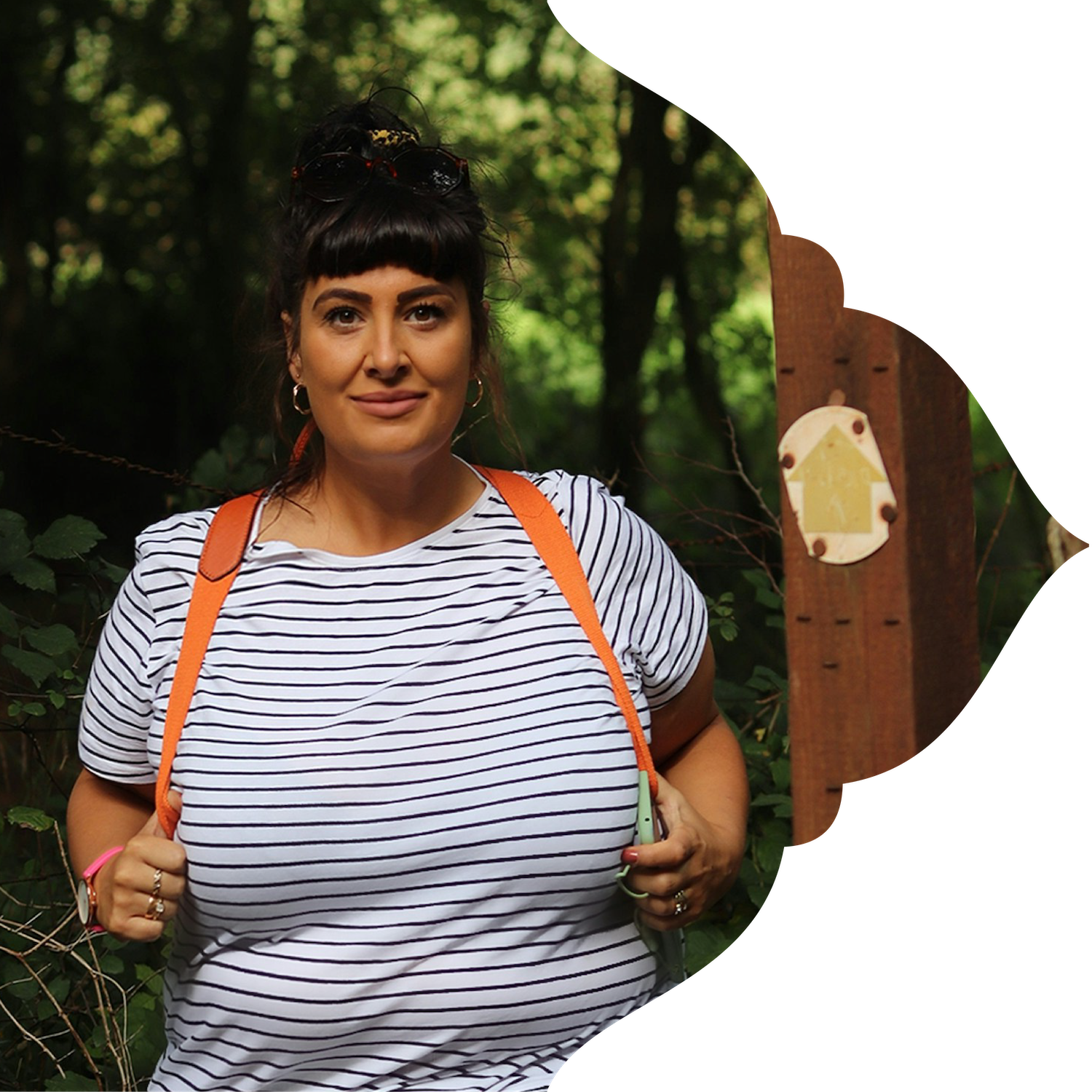 A woman with dark hair and bangs, wearing a white and navy striped shirt, stands outdoors in a wooded area, holding the straps of an orange backpack, with greenery in the background.