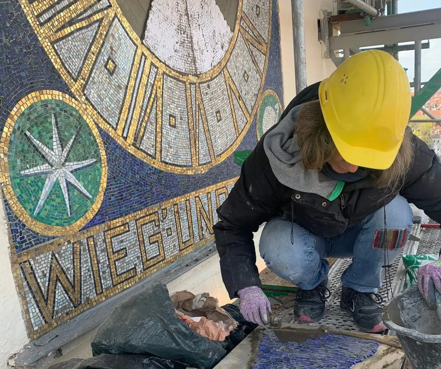 A worker wearing a yellow safety helmet and gloves installing a mosaic tile artwork on a wall. The mosaic includes clock faces and the word 'WIELGIN'.