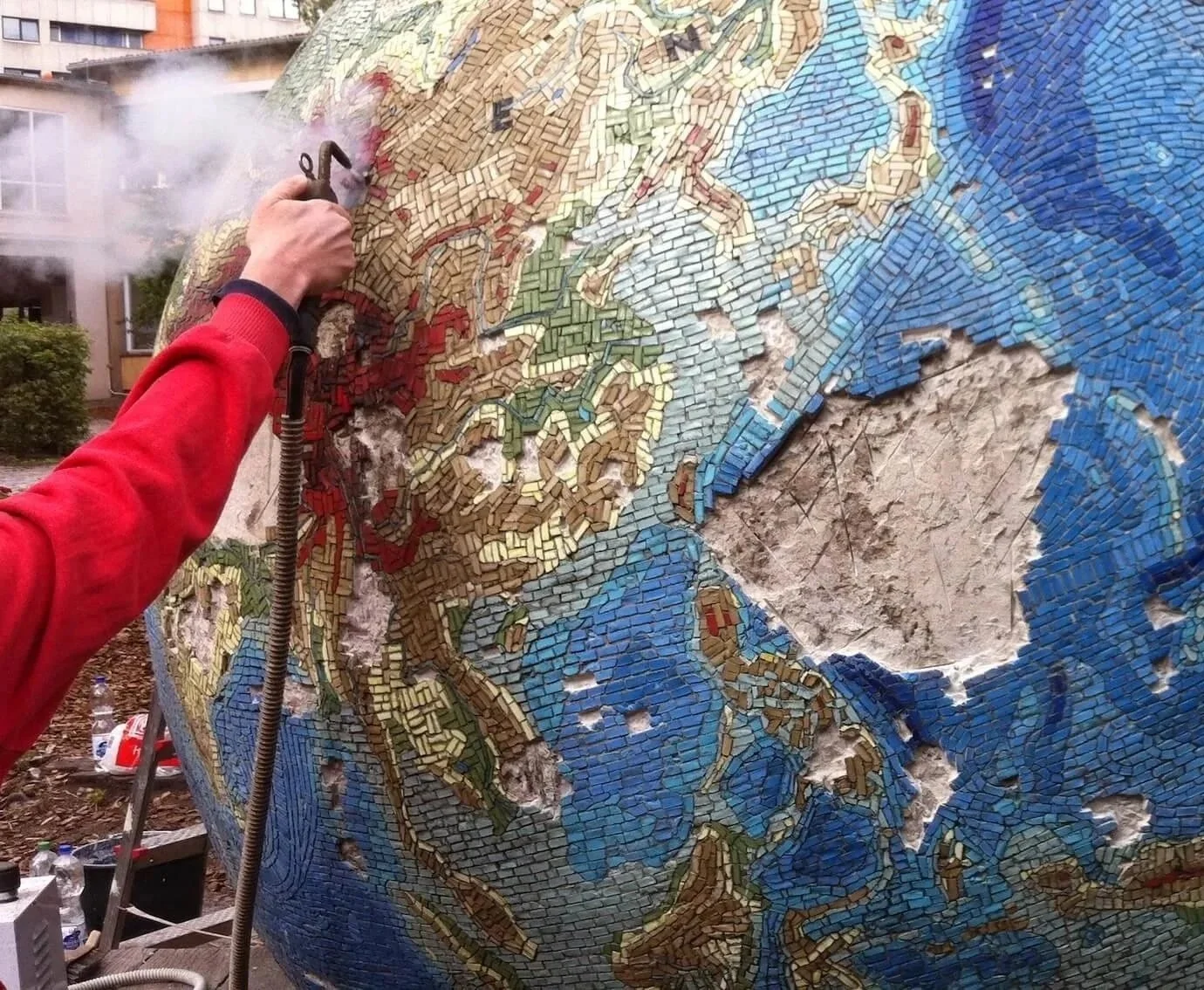 A person in a red jacket cleaning a large outdoor mosaic sculpture of the Earth with a power washer. The mosaic depicts continents and oceans using small, colorful tiles.