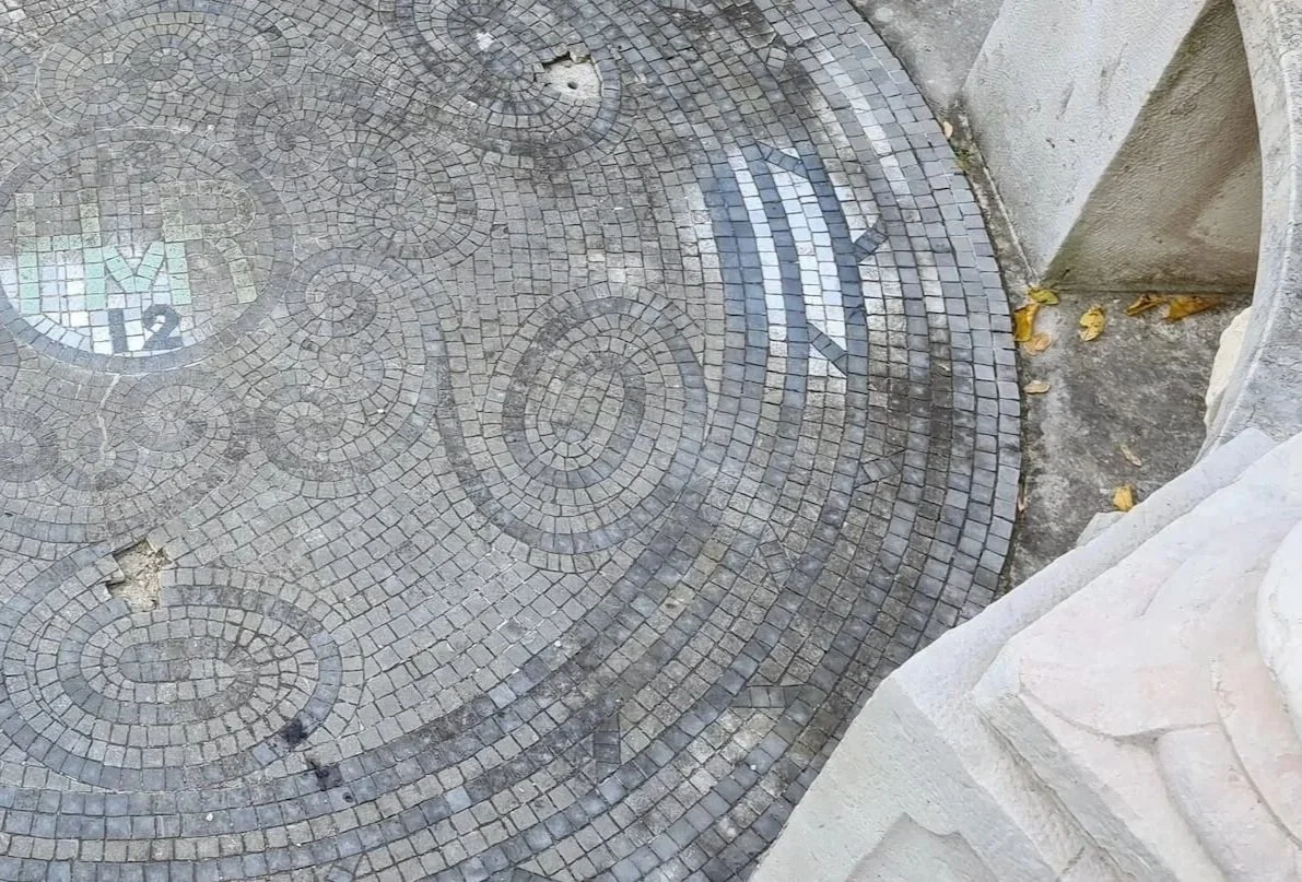 Decorative mosaic tile floor with spiral and fish designs outside, with a stone wall and scattered fallen leaves nearby.