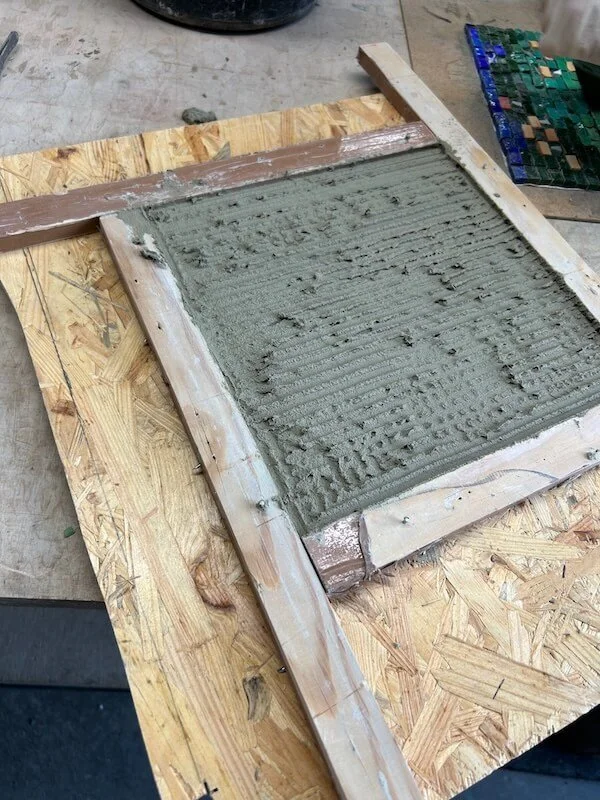 Screen frame with wet cement or concrete applied, resting on a wooden work surface.