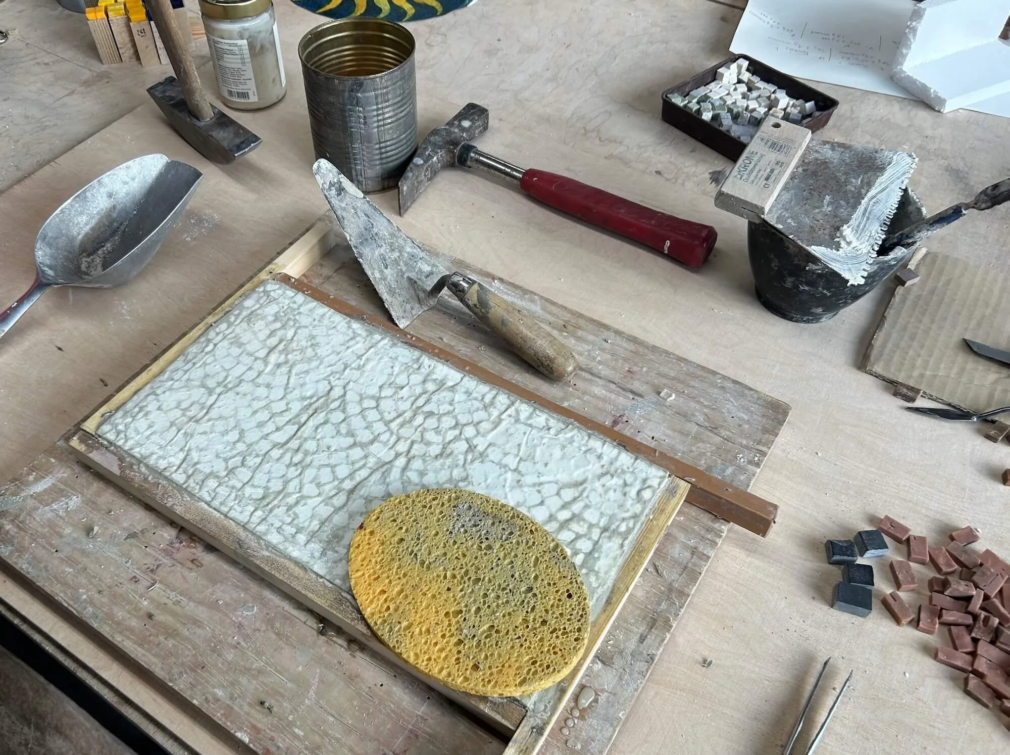 A workbench with a textured ceramic tile, a yellow sponge, various tools including a trowel, a chisel, and a hammer, along with containers of miscellaneous materials and small tiles.