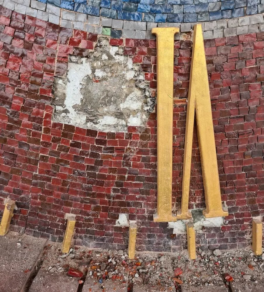 Close-up of a brick wall with an area of damaged or missing bricks and plaster. Five wooden support beams are leaning against the wall, likely for construction or repair work.