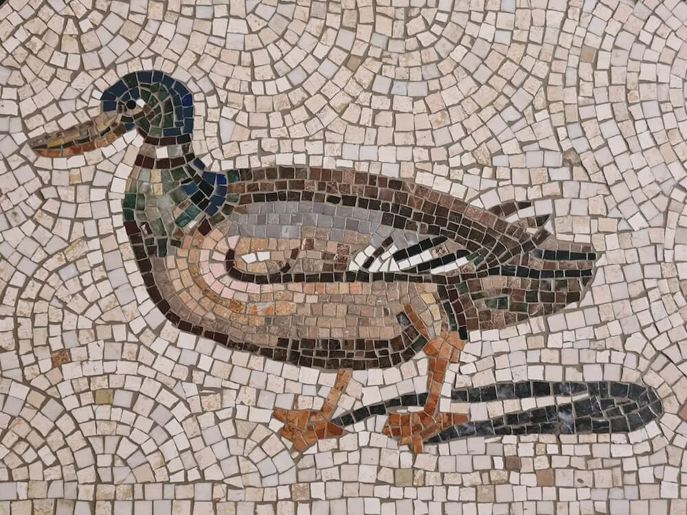 Mosaic of a duck with a black beak, brown body, and black tail on a beige tiled background.