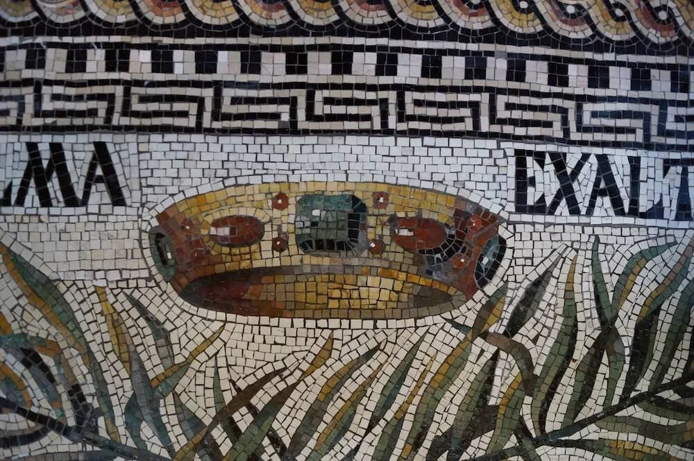 Close-up view of a mosaic with a circular emblem, Roman-style Roman numerals, and Greek key pattern at the top. The mosaic depicts a crown and decorative olive branches.