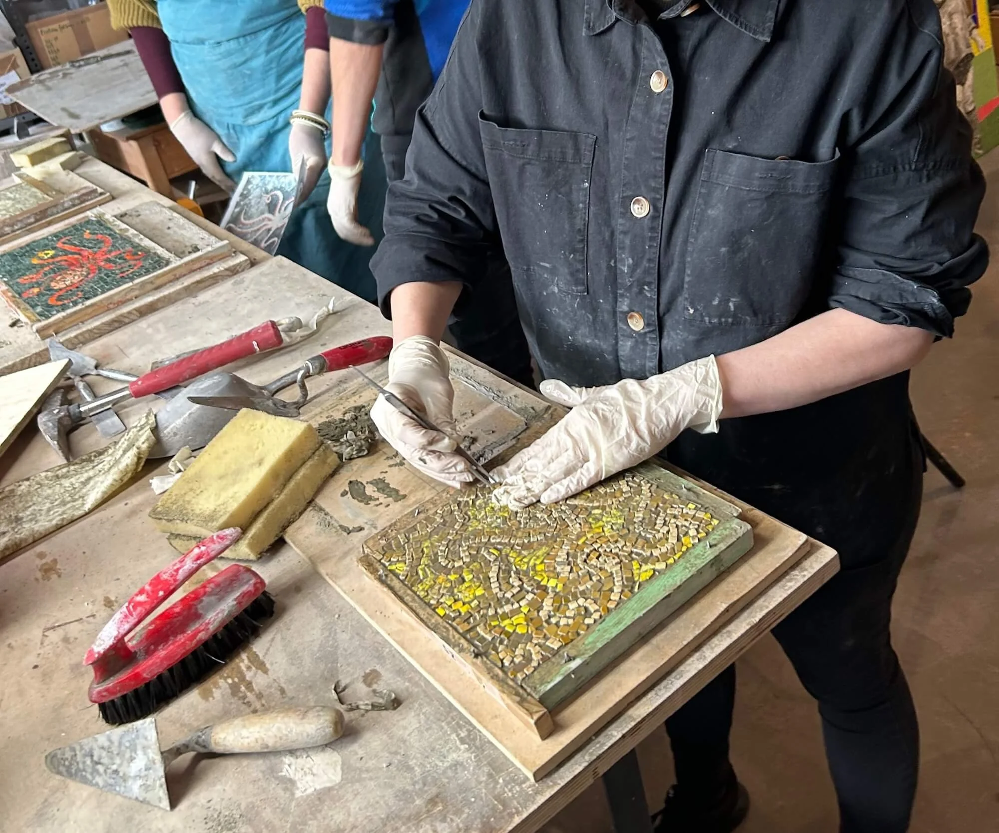 Person working on a mosaic artwork, wearing gloves, using tools on a worktable with various tiles and equipment, in an art studio.